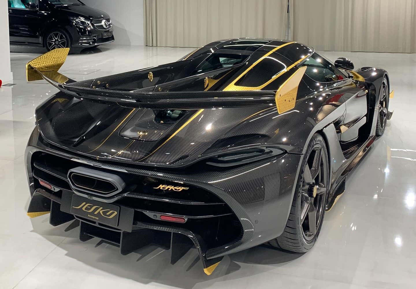  壁紙 — 一輛時尚的黑色和金色 Koenigsegg Jesko 超級跑車，採用碳纖維車身和大尾翼，在明亮的展廳中展示。