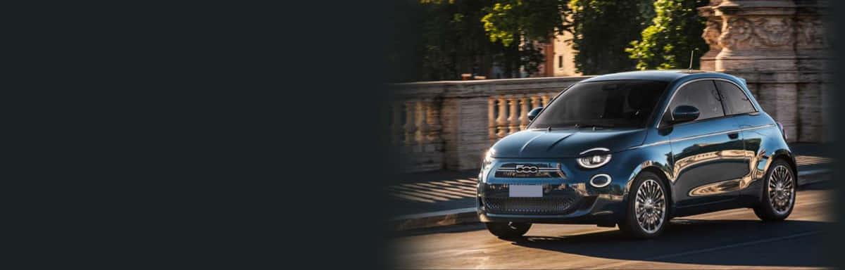 Fiat Panda 桌布 壁紙 — 一輛時尚的藍色Fiat 500e電動車在金色陽光下，在兩旁種滿樹木、擁有石頭建築的都市街道上閃閃發光。