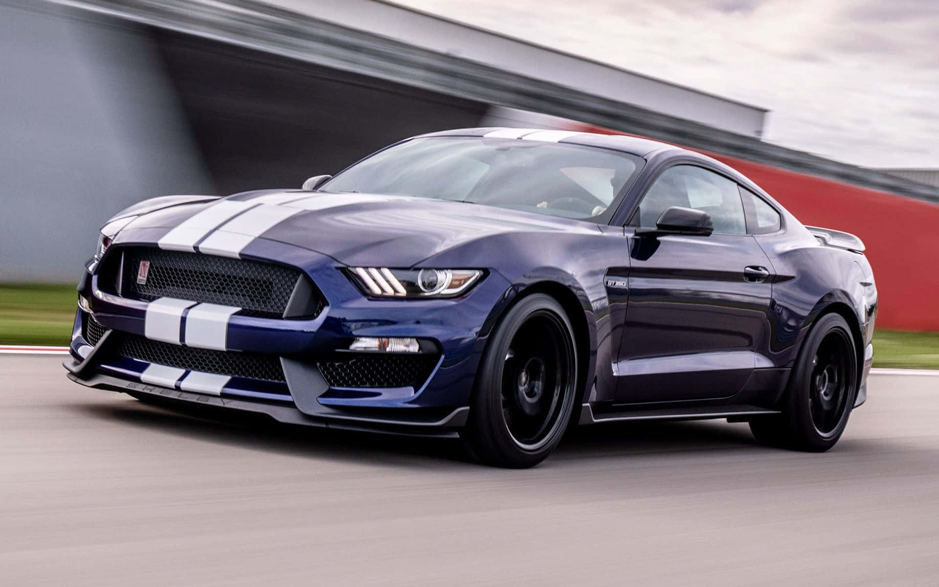 Ford Mustang Shelby Gt350 桌布 壁紙 — 一輛時尚的藍色福特野馬，帶有白色賽車條紋，在多雲的天空下沿著賽道飛馳。
