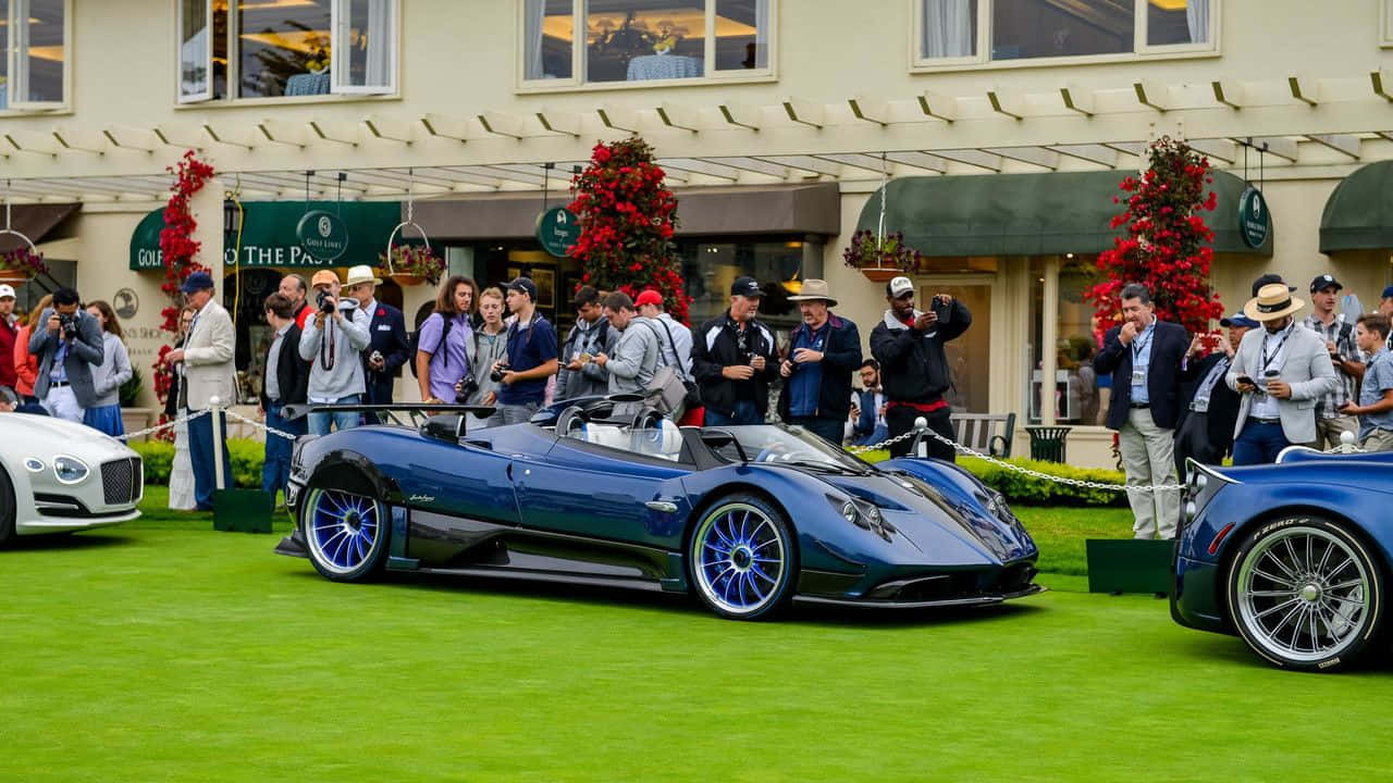 Pagani Zonda Hp Barchetta 桌布 壁紙 — 一輛時尚的藍色Pagani Huayra開篷跑車在修剪整齊的綠色草地上閃閃發光，攝影師和觀眾聚集在豪華車展活動中。