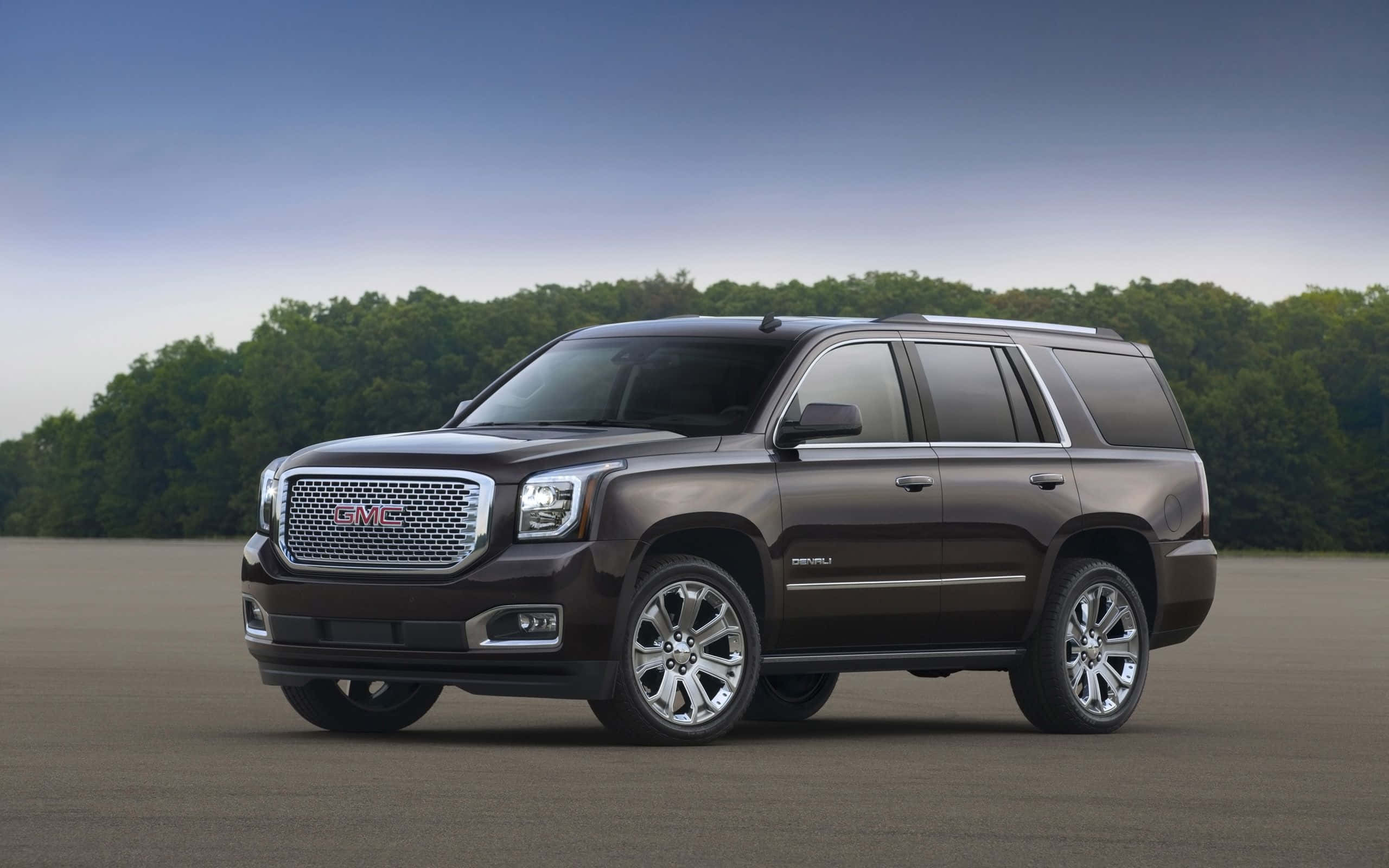 Gmc Yukon 桌布 壁紙 — 一輛時尚的深棕色GMC Yukon SUV停泊在鋪砌路面上，背景是晴朗藍天下的綠樹。