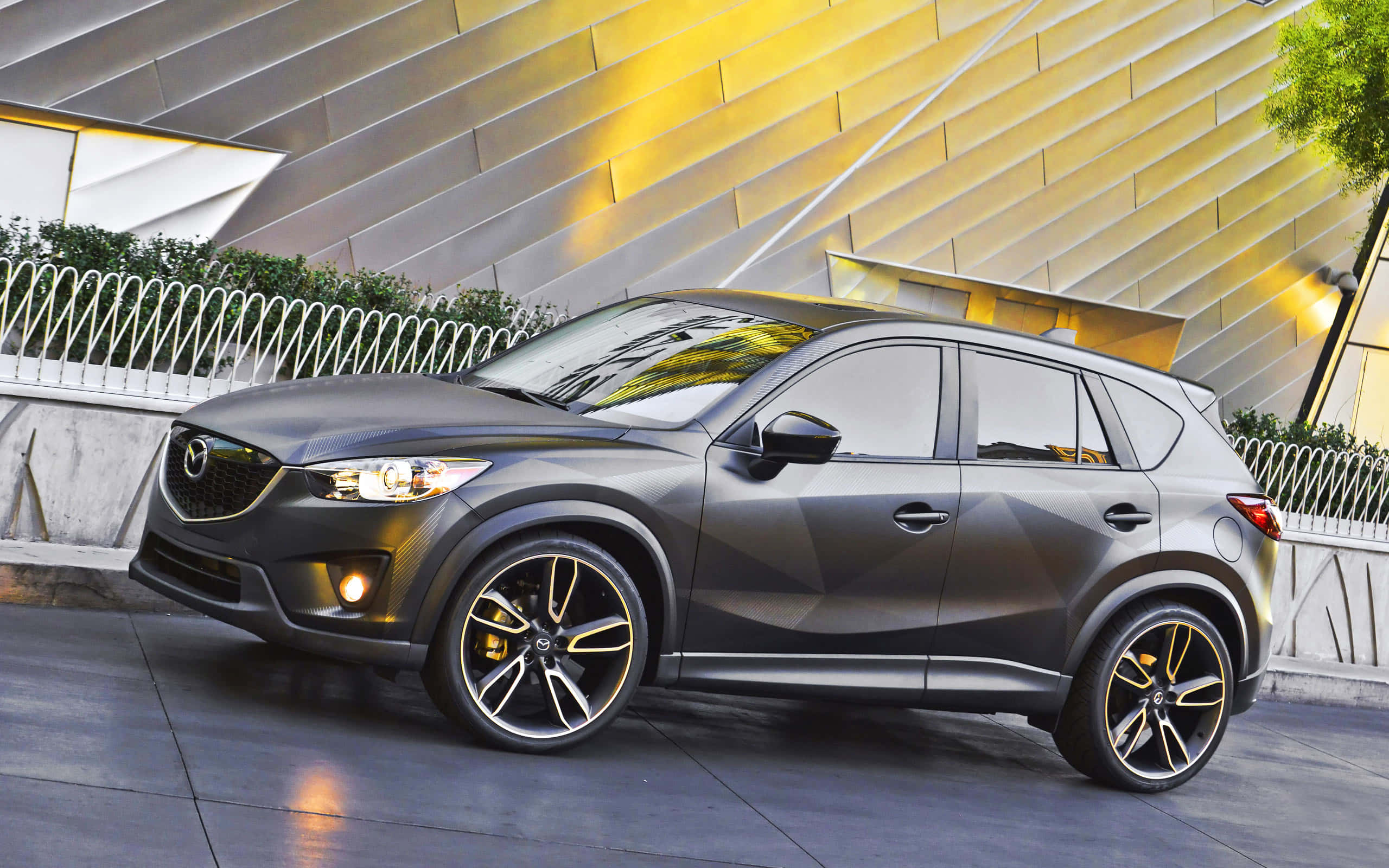 Mazda Cx-30 桌布 壁紙 — 一輛時尚的啞光灰色萬事得CX-5 SUV，配備金色輪圈，在黃昏時停在一座現代幾何建築前。
