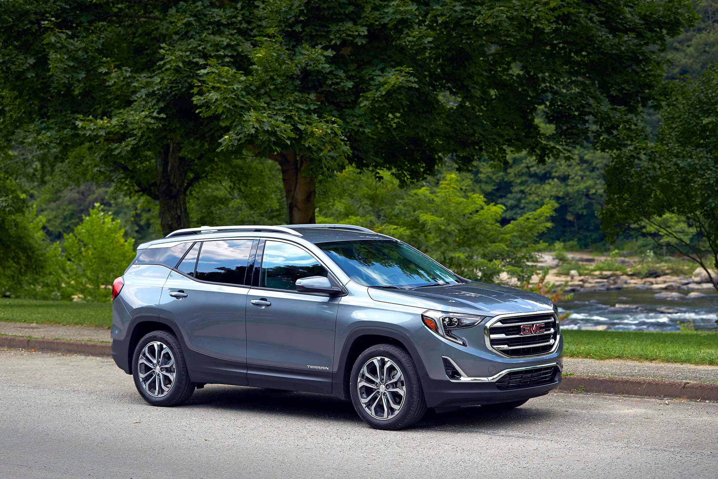 Gmc Terrain 桌布 壁紙 — 一輛時尚的銀色GMC Terrain SUV停泊在路邊，背景是茂密的綠樹和流淌的河流。