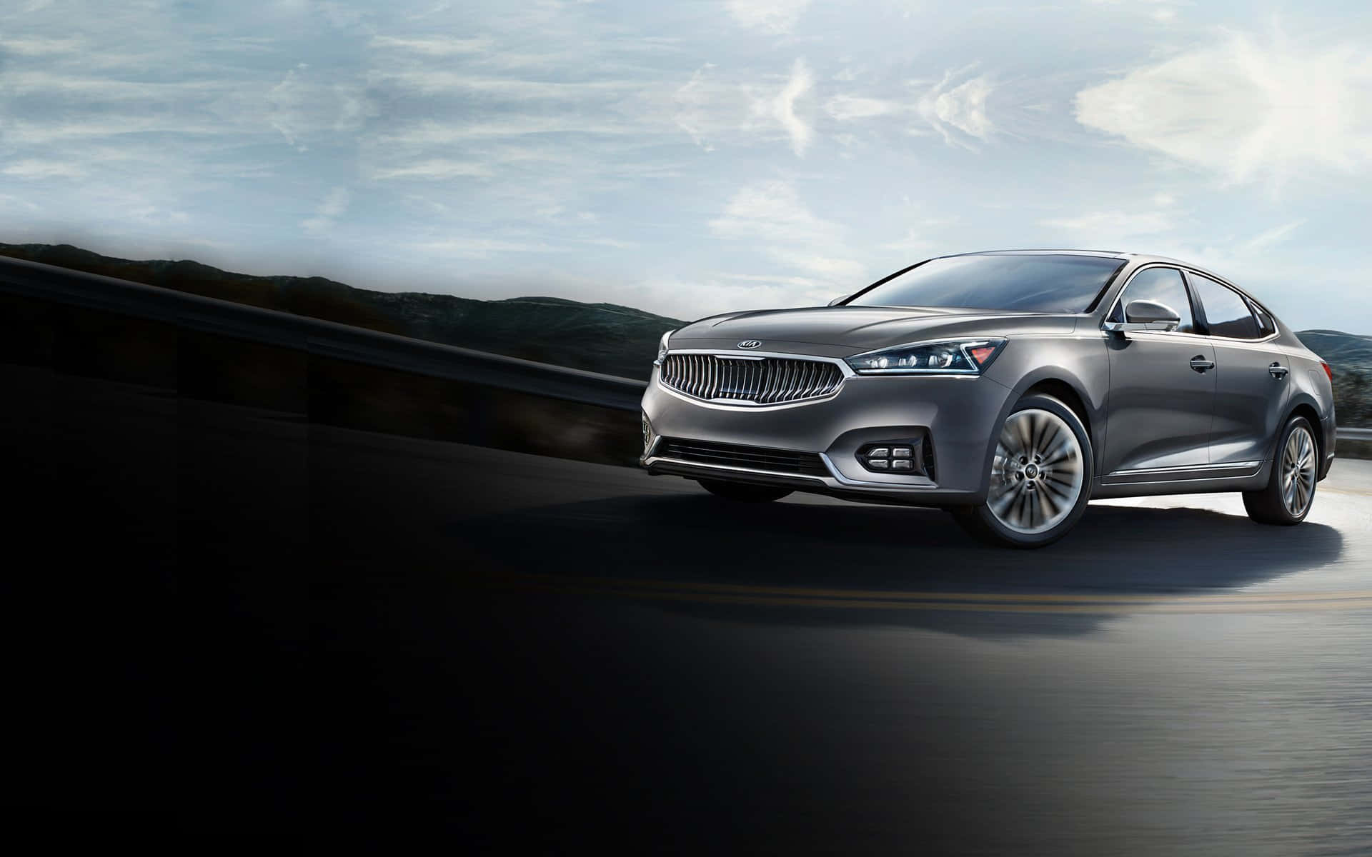 Kia Cadenza 桌布 壁紙 — 一輛時尚的銀色轎車在多雲的天空下沿著蜿蜒的山路疾馳。