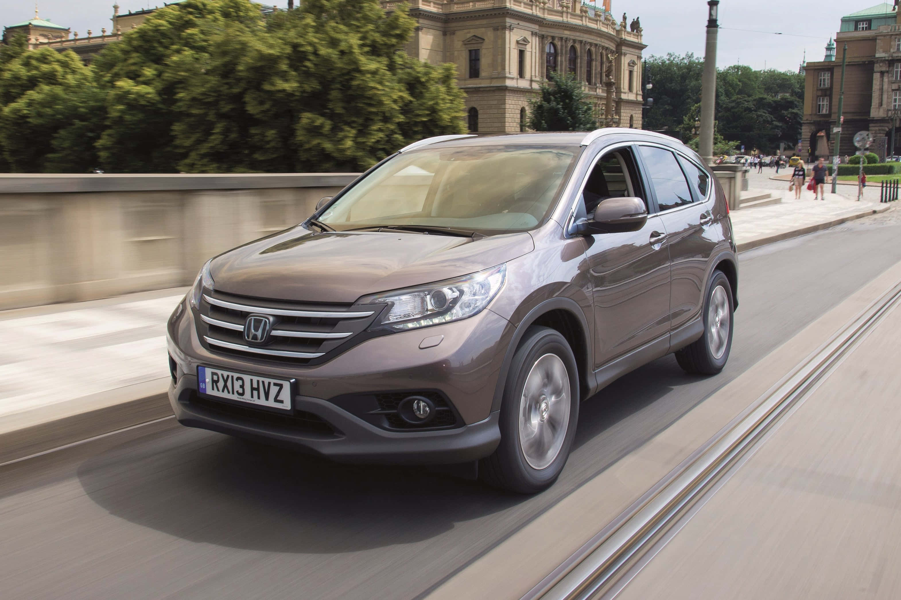 Honda Cr-v 桌布 壁紙 — 一輛時尚的棕色本田CR-V沿著城市街道行駛，背景是歷史建築和綠樹。