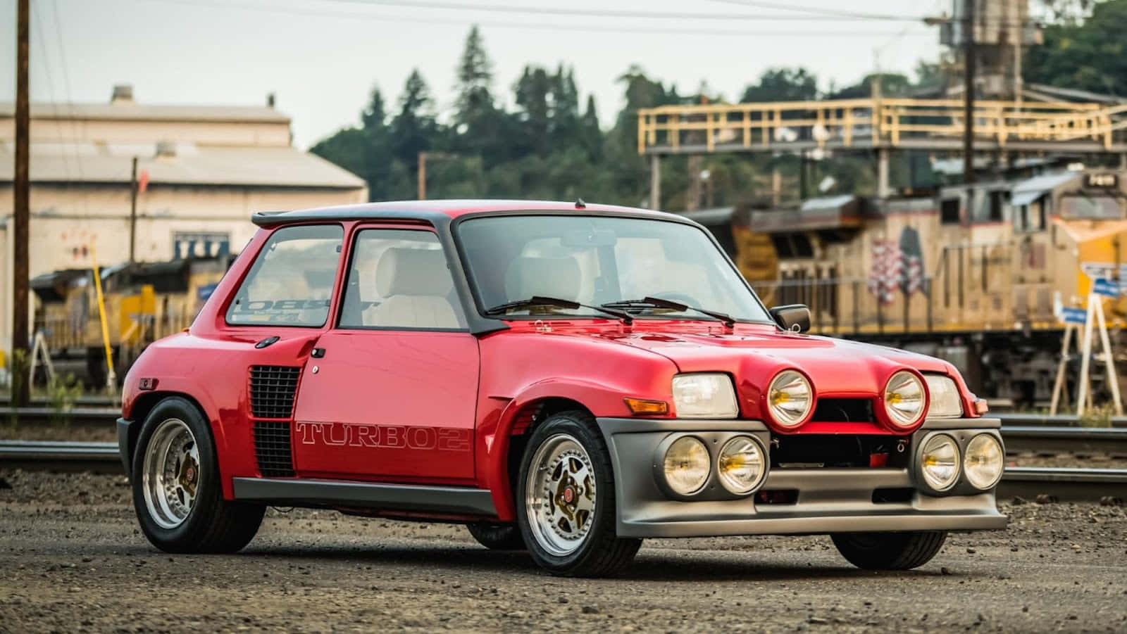 Renault 5 Turbo 桌布 壁紙 — 一輛鮮紅色的雷諾5 Turbo自豪地停在鐵軌旁的礫石上，背景有工業建築和樹木。