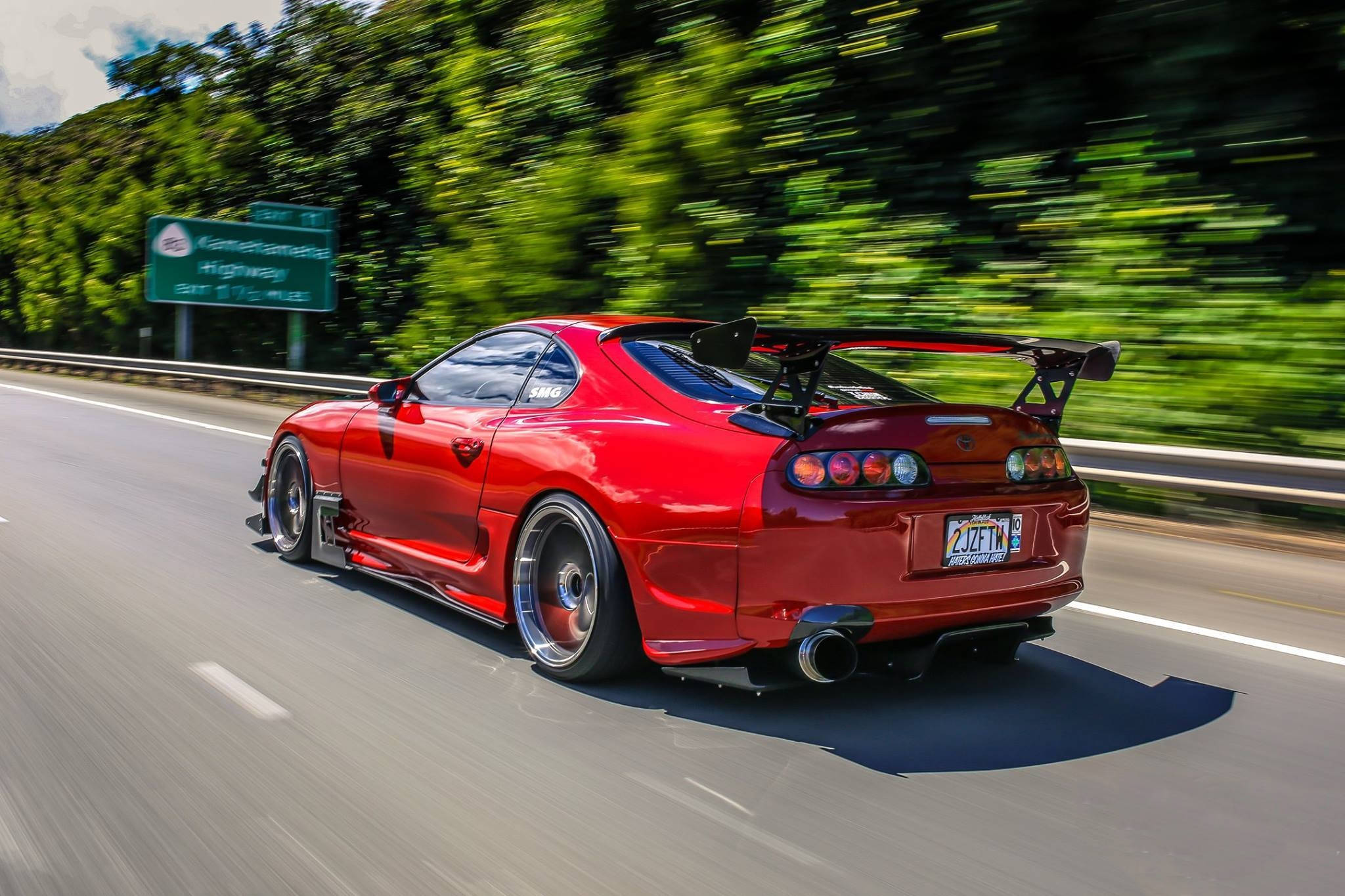 1998 年 Toyota Supra Mk4 行駛於高速公路 壁紙 — 一輛鮮紅色的跑車在陽光明媚的高速公路上疾馳，茂密的綠樹在運動中模糊成影。