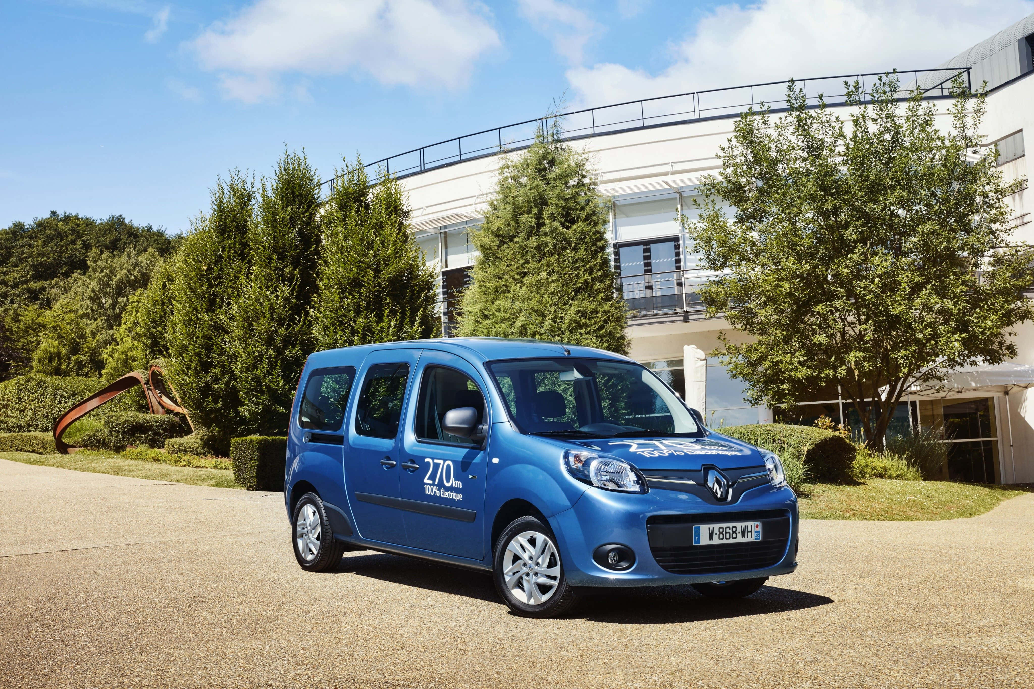 Renault Kangoo 桌布 壁紙 — 一輛鮮藍色的雷諾Kangoo停泊在陽光普照的車道上，旁邊有綠樹和現代建築。