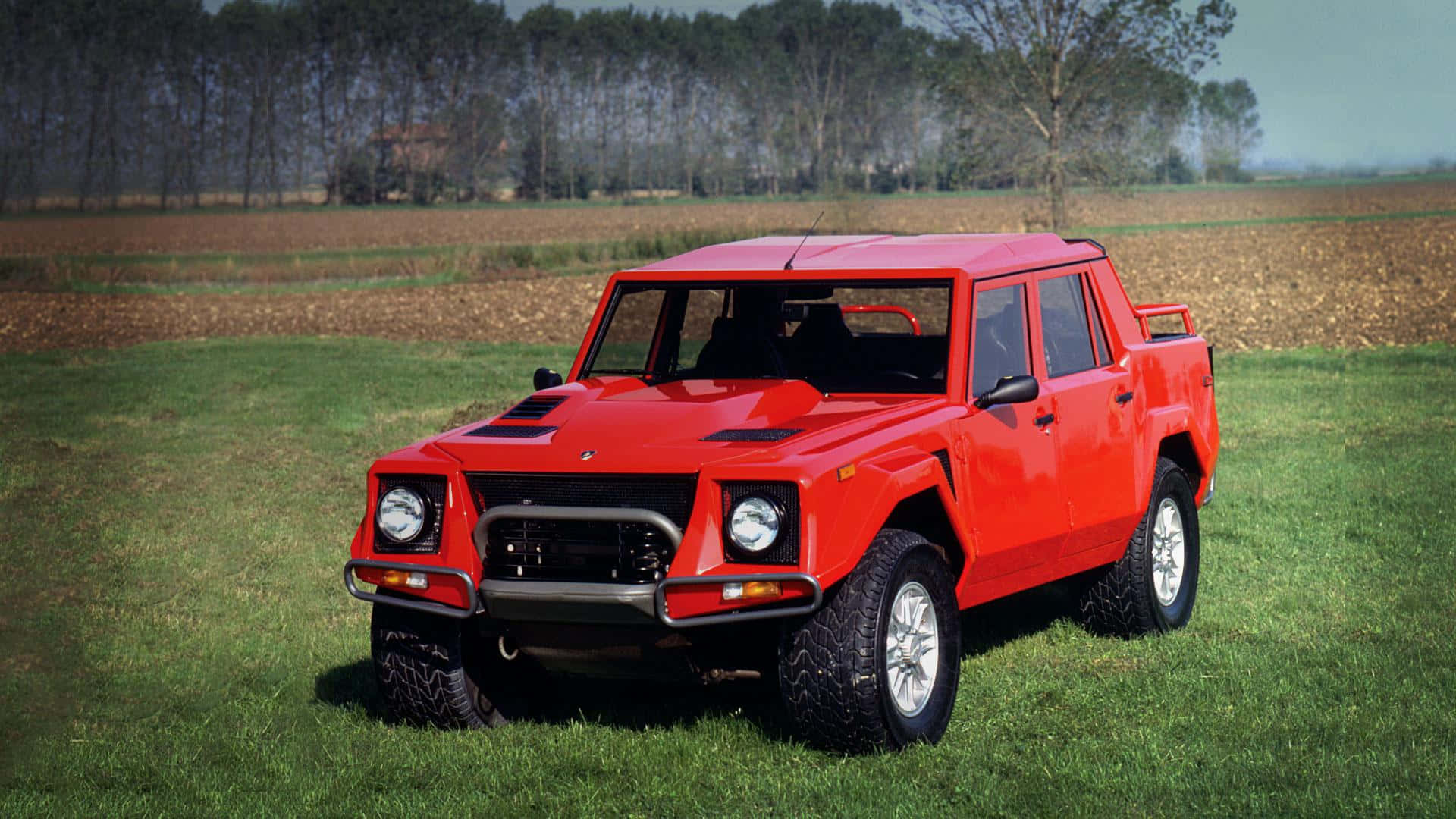 Lamborghini Lm002 桌布 壁紙 — 一輛醒目的紅色林寶堅尼LM002大膽地停在草地上，背景是樹木和農田，天空晴朗。