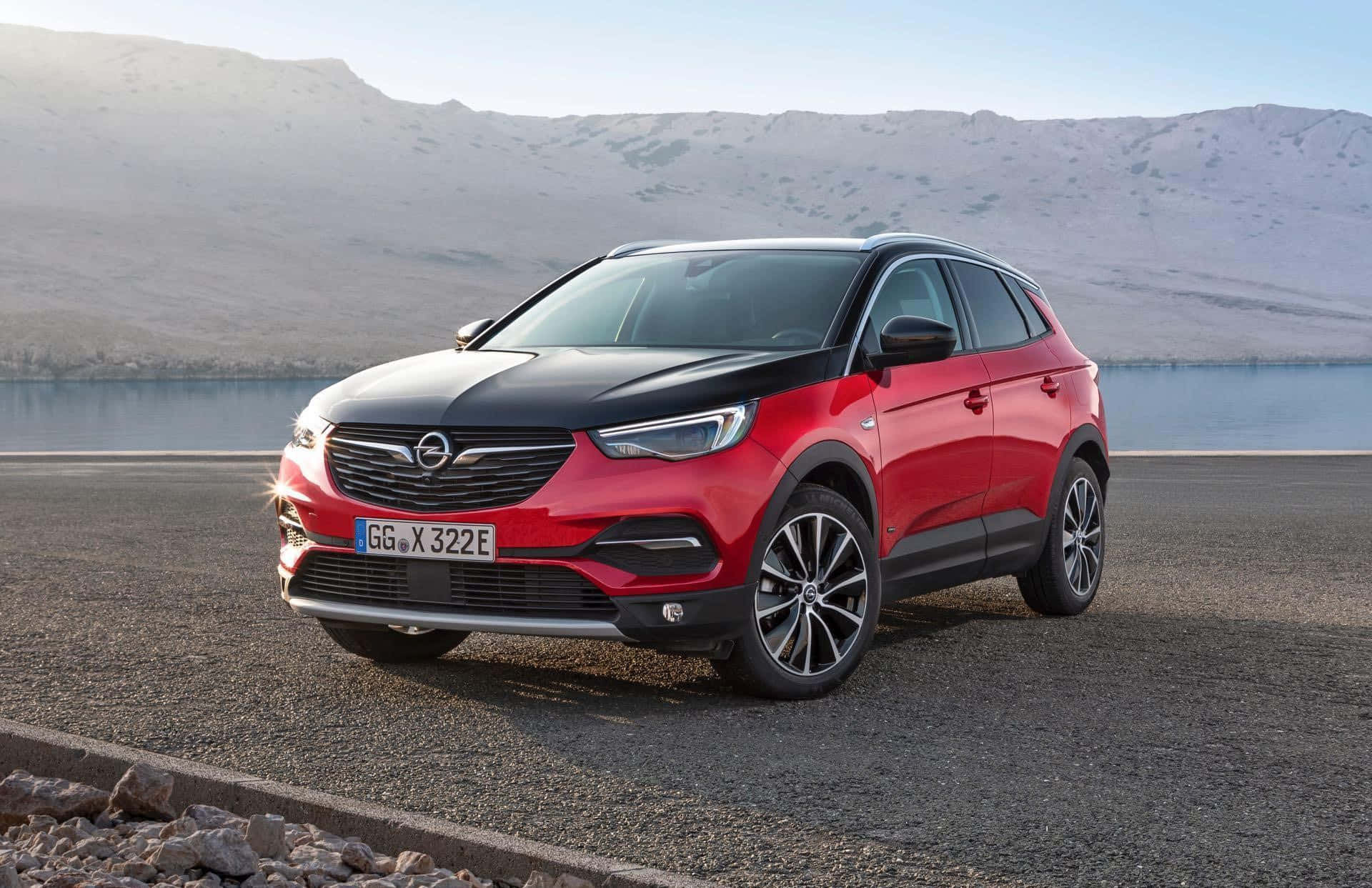 Opel Grandland X 桌布 壁紙 — 一輛醒目的紅色和黑色Opel Crossland X SUV停泊在碎石路上，旁邊是一個平靜的湖泊，背景是白雪覆蓋的山脈。