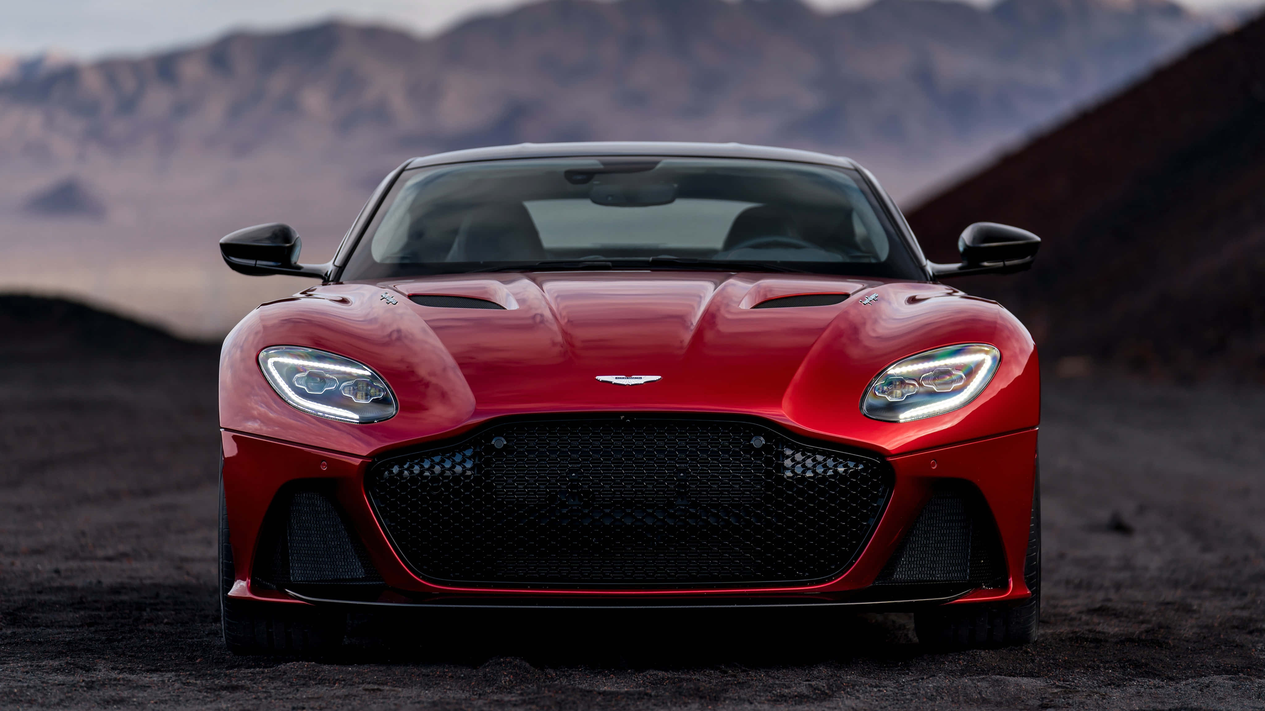 Aston Martin Dbs Superleggera 桌布 壁紙 — 一輛引人注目的紅色Aston Martin跑車在崎嶇的沙漠道路上向前行駛，背景是朦朧的山脈。