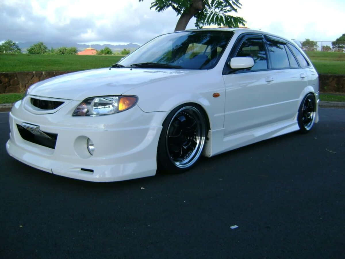 Mazda Protege 桌布 壁紙 — 一輛純白色改裝旅行車，黑色輪圈，停在瀝青路上，天空多雲，背景有綠草和樹木。
