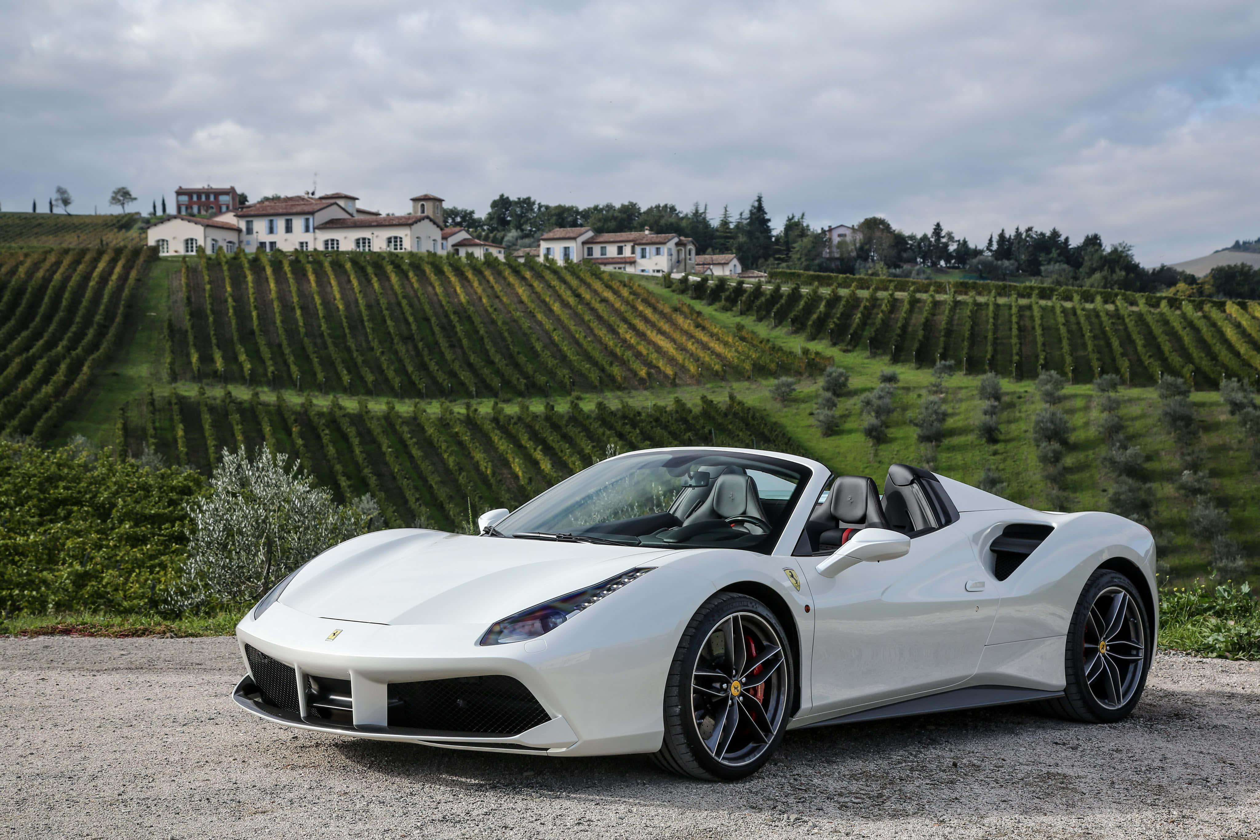 Ferrari 488 Spider 桌布 壁紙 — 一輛純白色的法拉利Spider停在碎石路上，背景是起伏的葡萄園和托斯卡納別墅，天空多雲。