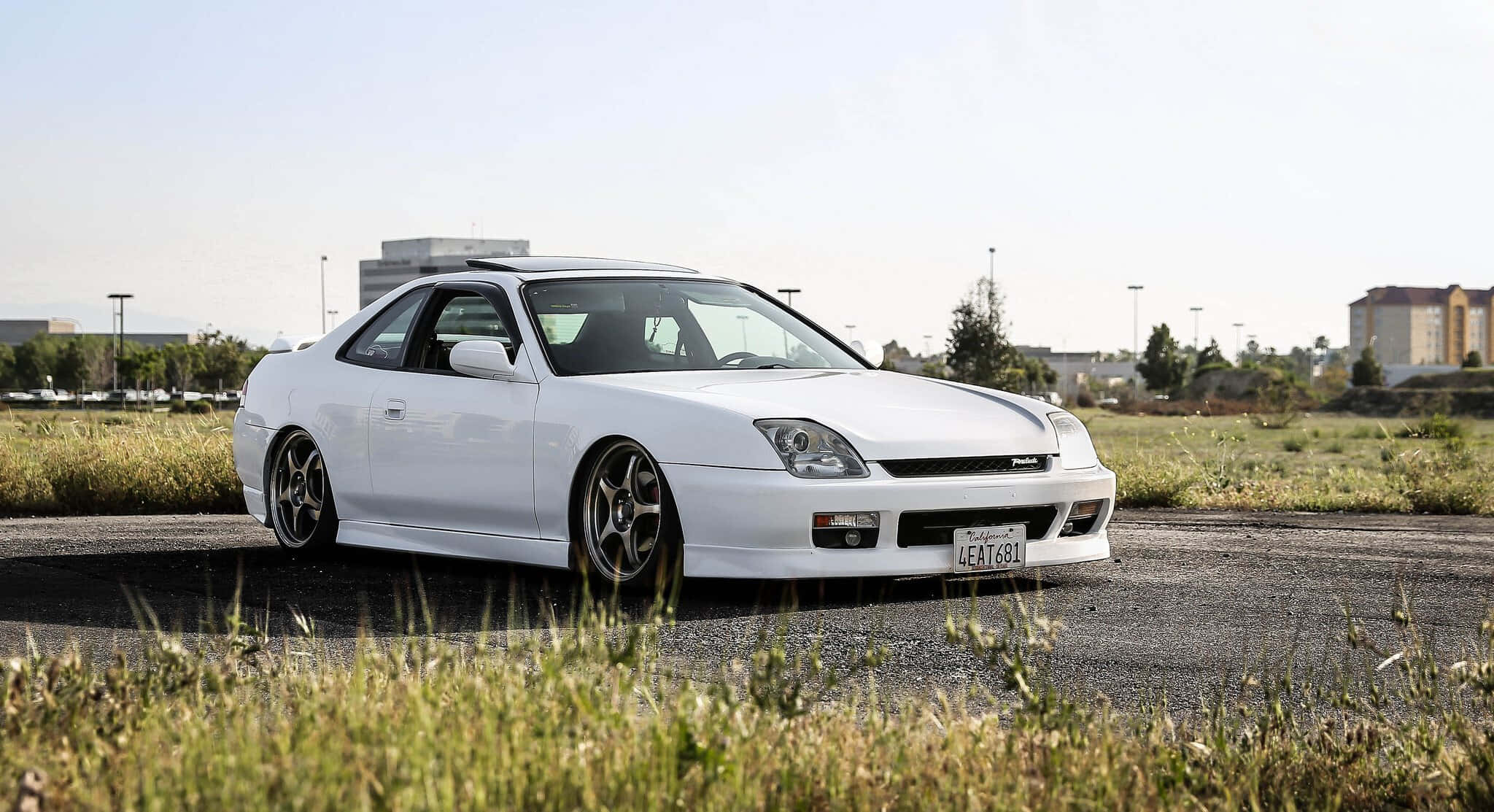 Honda Prelude 桌布 壁紙 — 一輛純白色運動型轎跑車配備定制輪圈，停在柏油路上，周圍是高高的草叢，天空明亮。