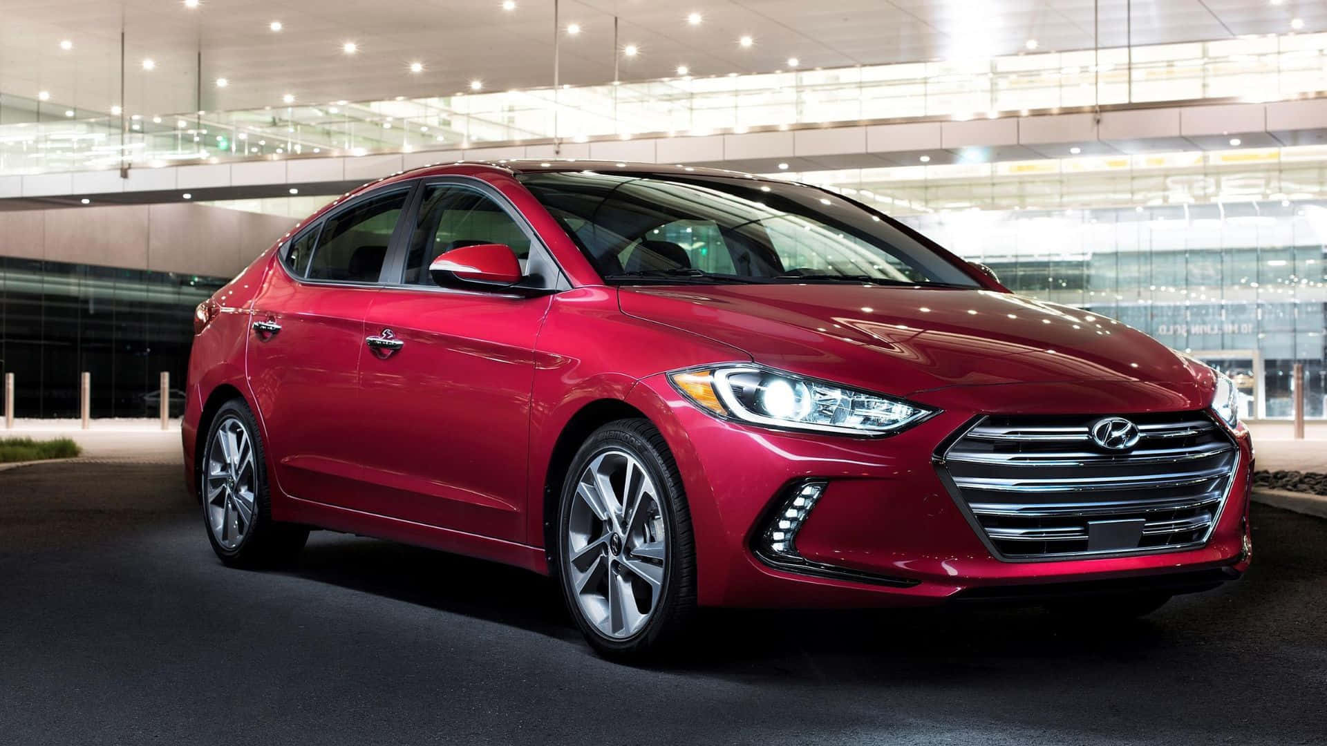 Hyundai Elantra 桌布 壁紙 — 一輛光亮的紅色現代房車在現代玻璃建築停車區的明亮燈光下閃閃發光。