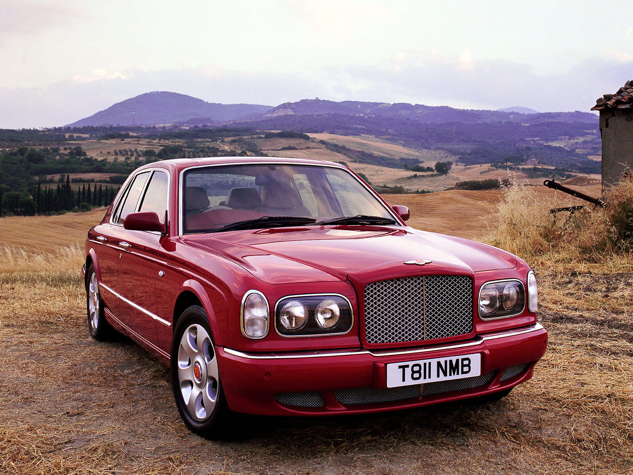 Bentley Arnage 桌布 壁紙 — 一輛光亮的紅色賓利房車停在乾草地上，背景有起伏的山丘和一座石建築，天空多雲。
