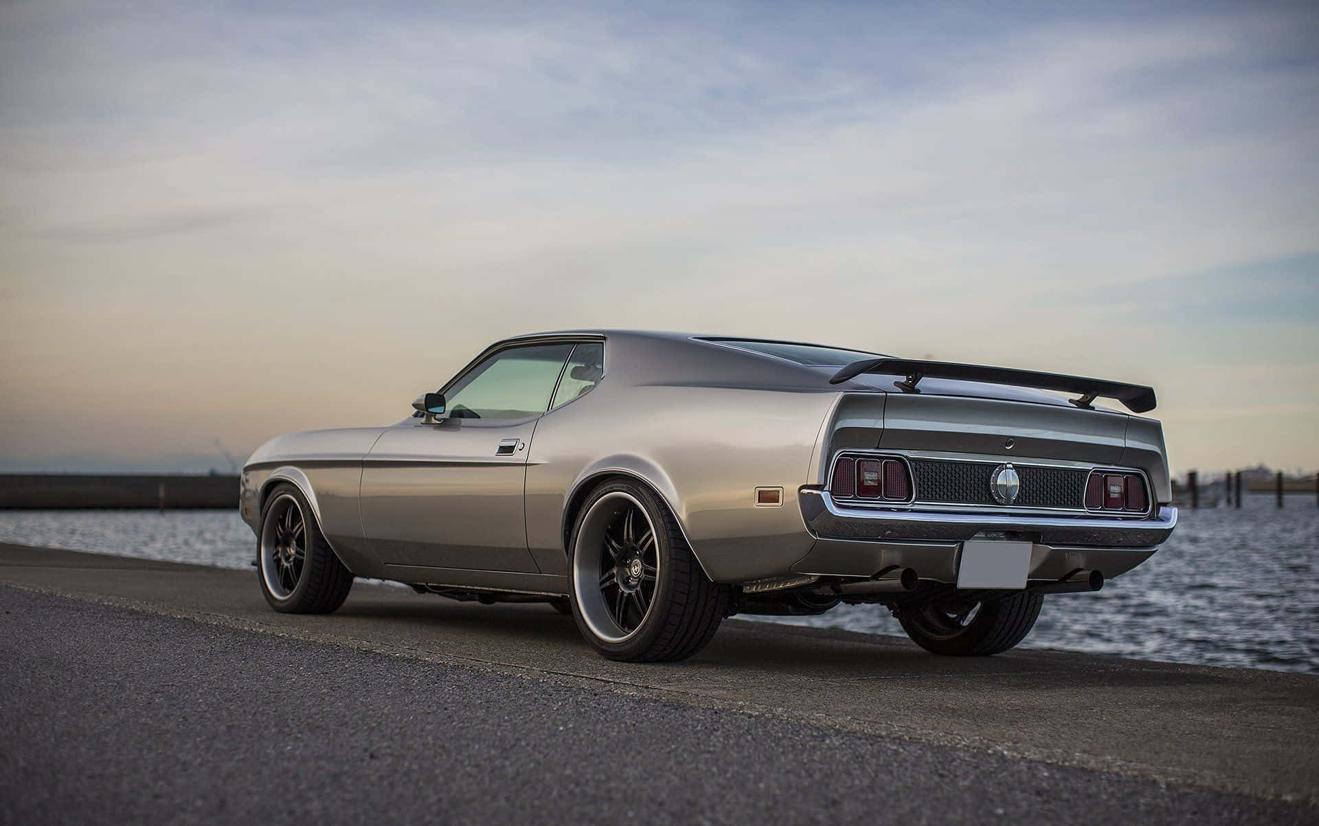 Ford Mustang Mach 1 桌布 壁紙 — 一輛銀色經典肌肉車，帶有黑色尾翼，停泊在日落時分的海岸公路上。