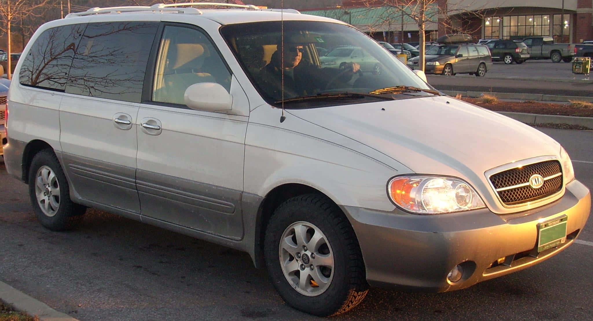 Kia Sedona 桌布 壁紙 — 一輛銀色Kia小型貨車停泊在一個停車場，背景有光禿禿的樹木和其他車輛。