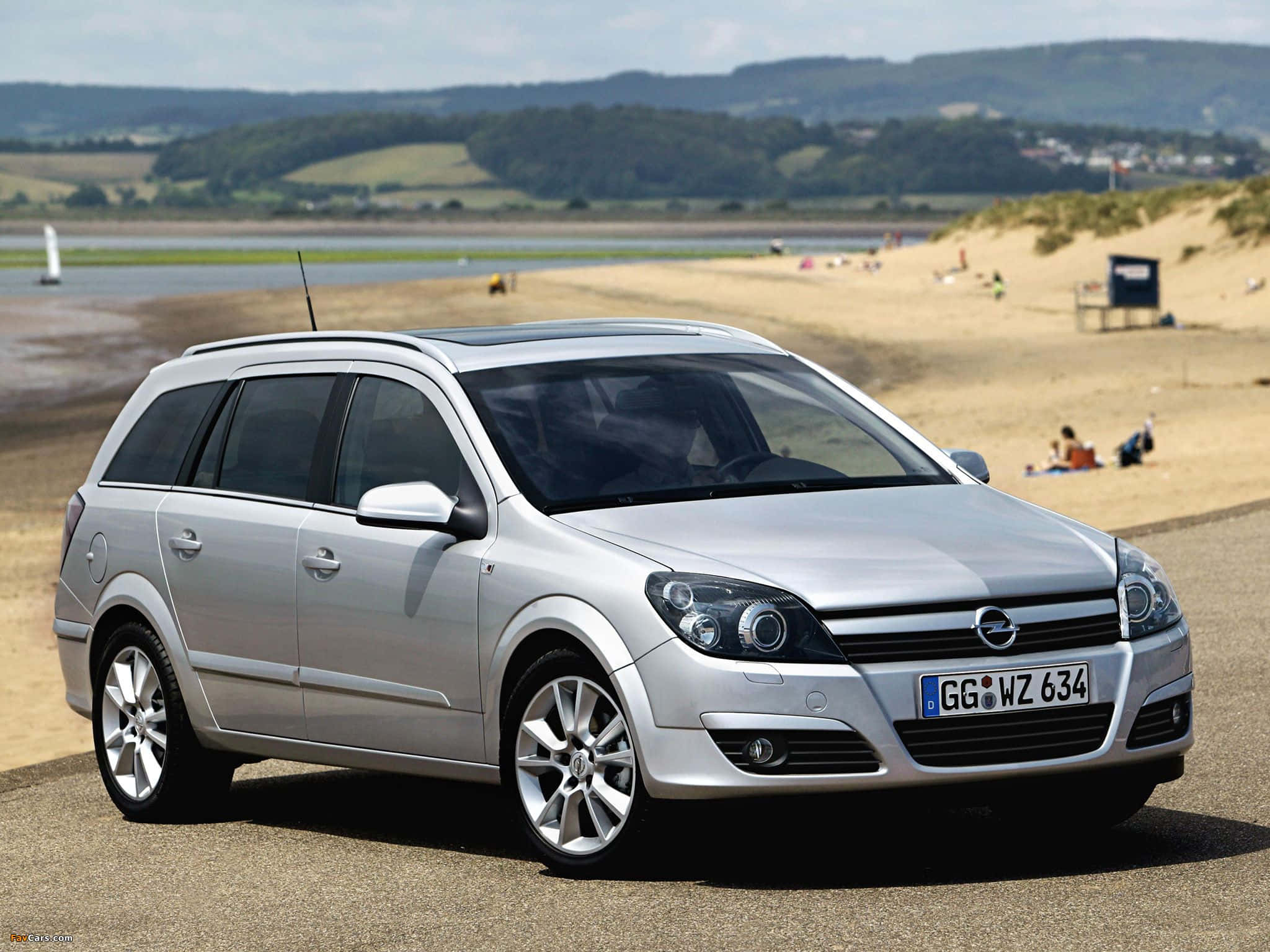 Opel Astra 桌布 壁紙 — 一輛銀色歐寶旅行車停泊在沙灘旁的碎石路上，背景有放鬆的人們和山丘。