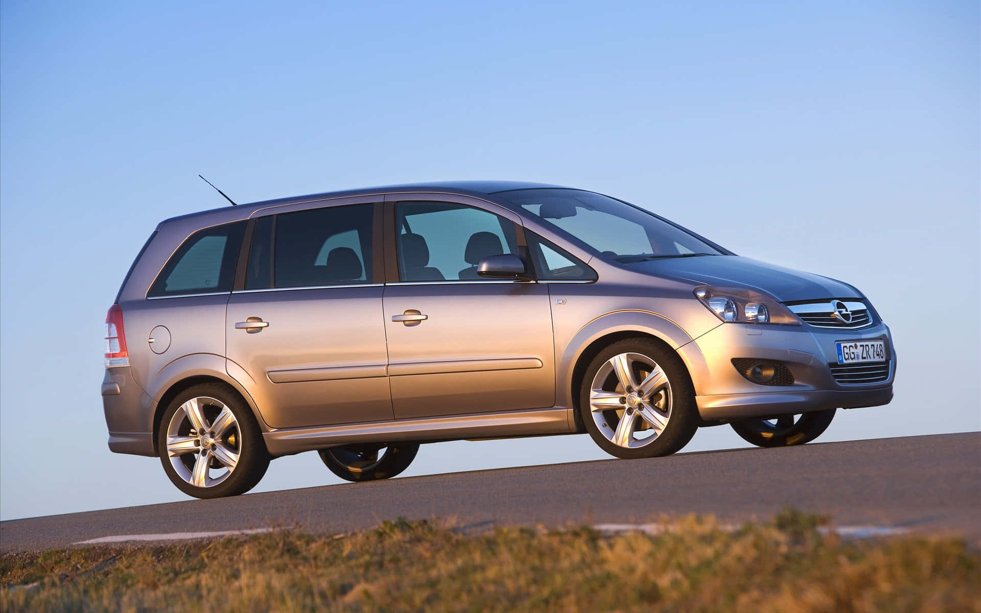 Opel Zafira 桌布 壁紙 — 一輛銀色歐寶Zafira旅行車在晴朗藍天下沿著鋪砌道路行駛，下方是金色草地。