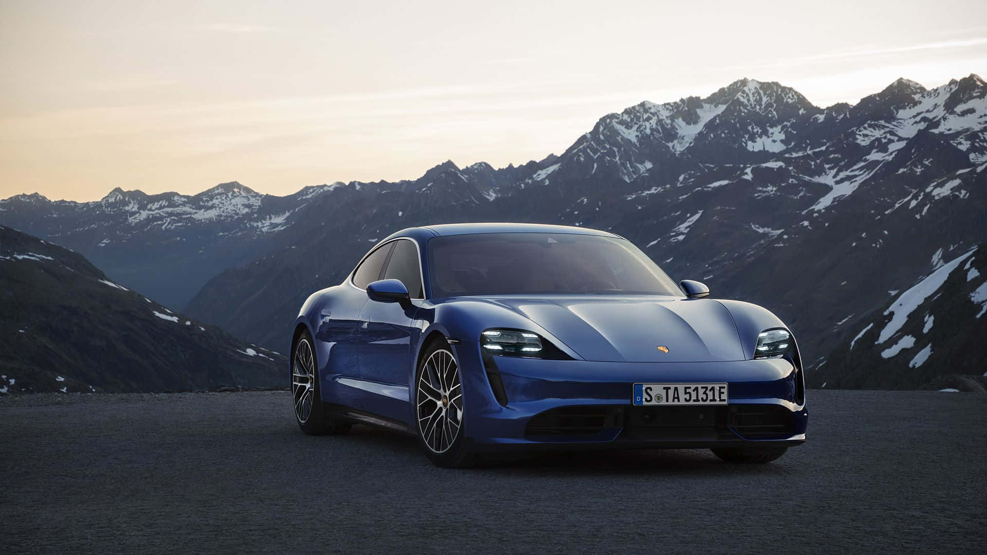 Porsche Taycan 桌布 壁紙 — 一輛時尚的藍色電動跑車停在山路上，背景是白雪覆蓋的山峰，天空呈現柔和的金色日落。