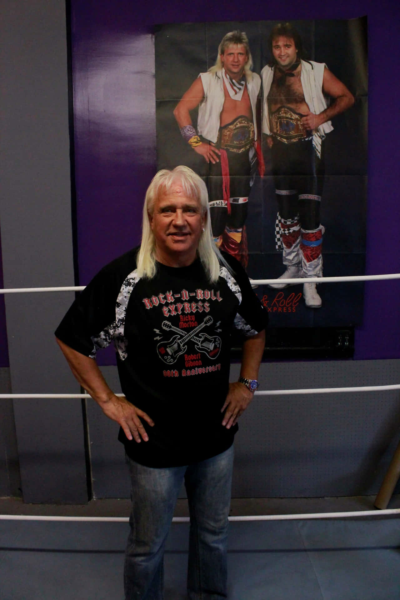  壁紙 — 一名長白髮的男子自信地站在摔角擂台上，身穿Rock n Roll Express T恤，身後有二人組的海報。