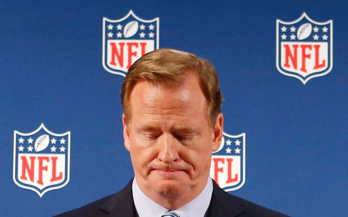  壁紙 — 一名穿西裝的男子在重複NFL標誌的藍色背景下，帶著陰沉的表情向下看。