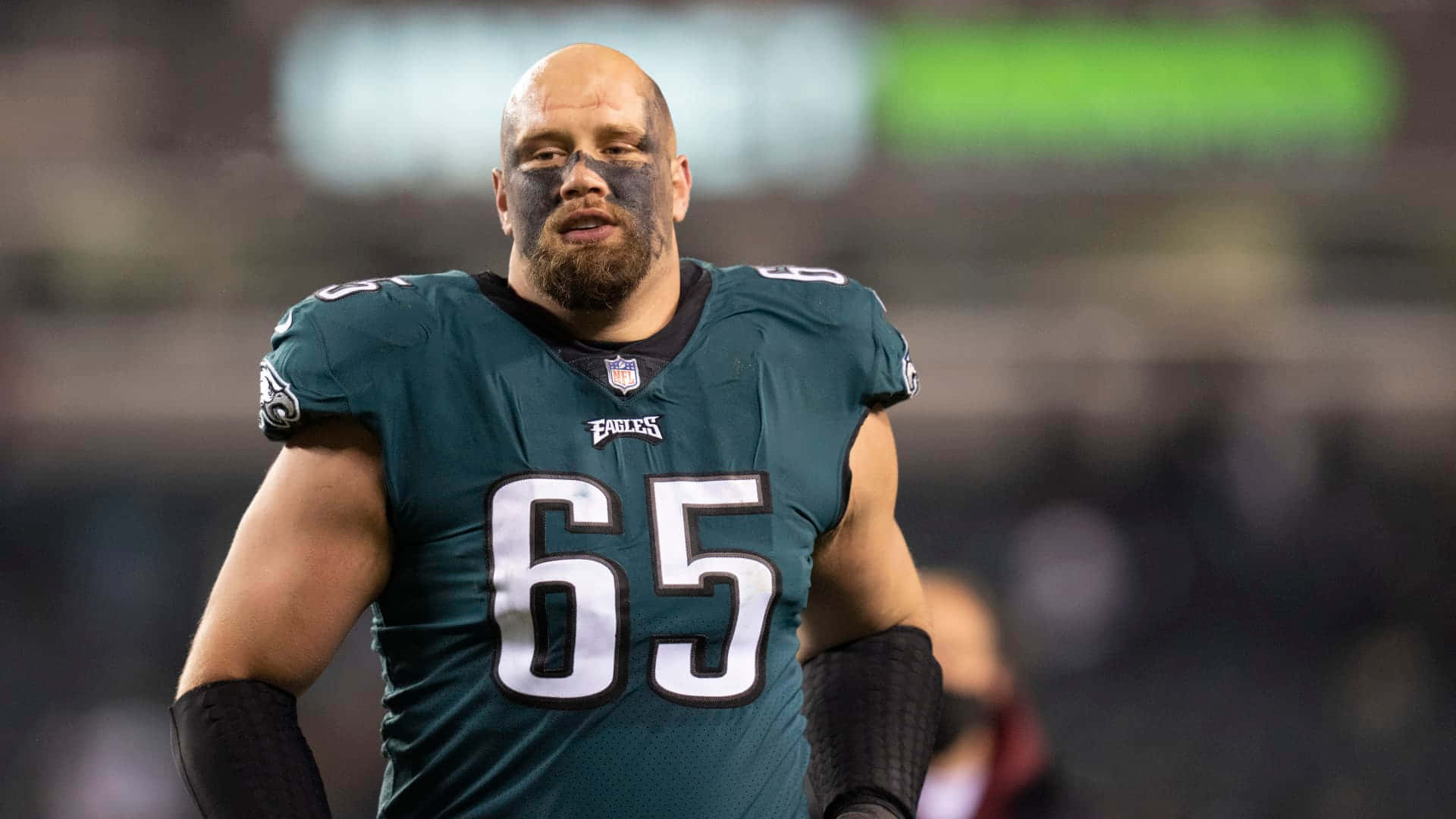  壁紙 — 一名光頭NFL進攻截鋒，臉上塗有黑色顏料並留鬍鬚，在夜間比賽中穿著綠色老鷹隊65號球衣。