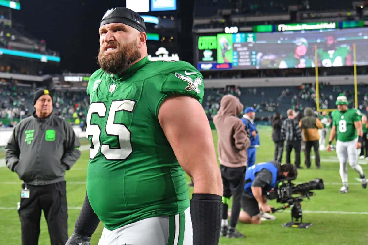  壁紙 — 一名肌肉發達的NFL進攻鋒線球員身穿綠色球衣，在夜間比賽中站在燈光璀璨的足球場上，背景有體育場燈光和記分牌。