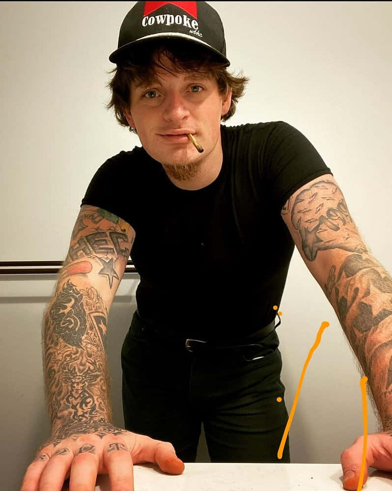  壁紙 — 一名紋身男子戴著黑色 'Cowpoke' 帽子和襯衫，嘴裡叼著香煙，靠在一面素牆上向前傾。