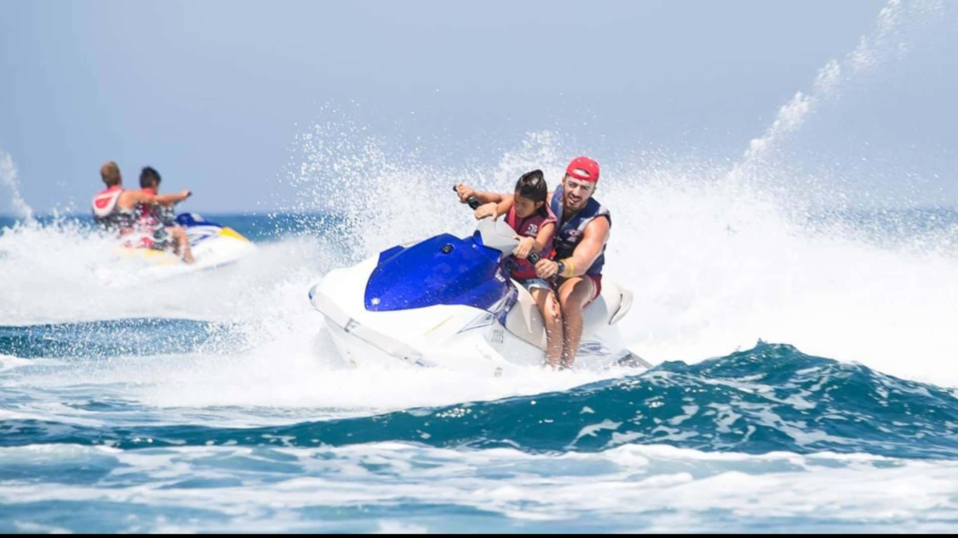 Beach Water Sports 桌布 壁紙 — 一名男子和一個小孩騎著藍白色的水上電單車，在清澈藍天下穿越碧綠的海浪。