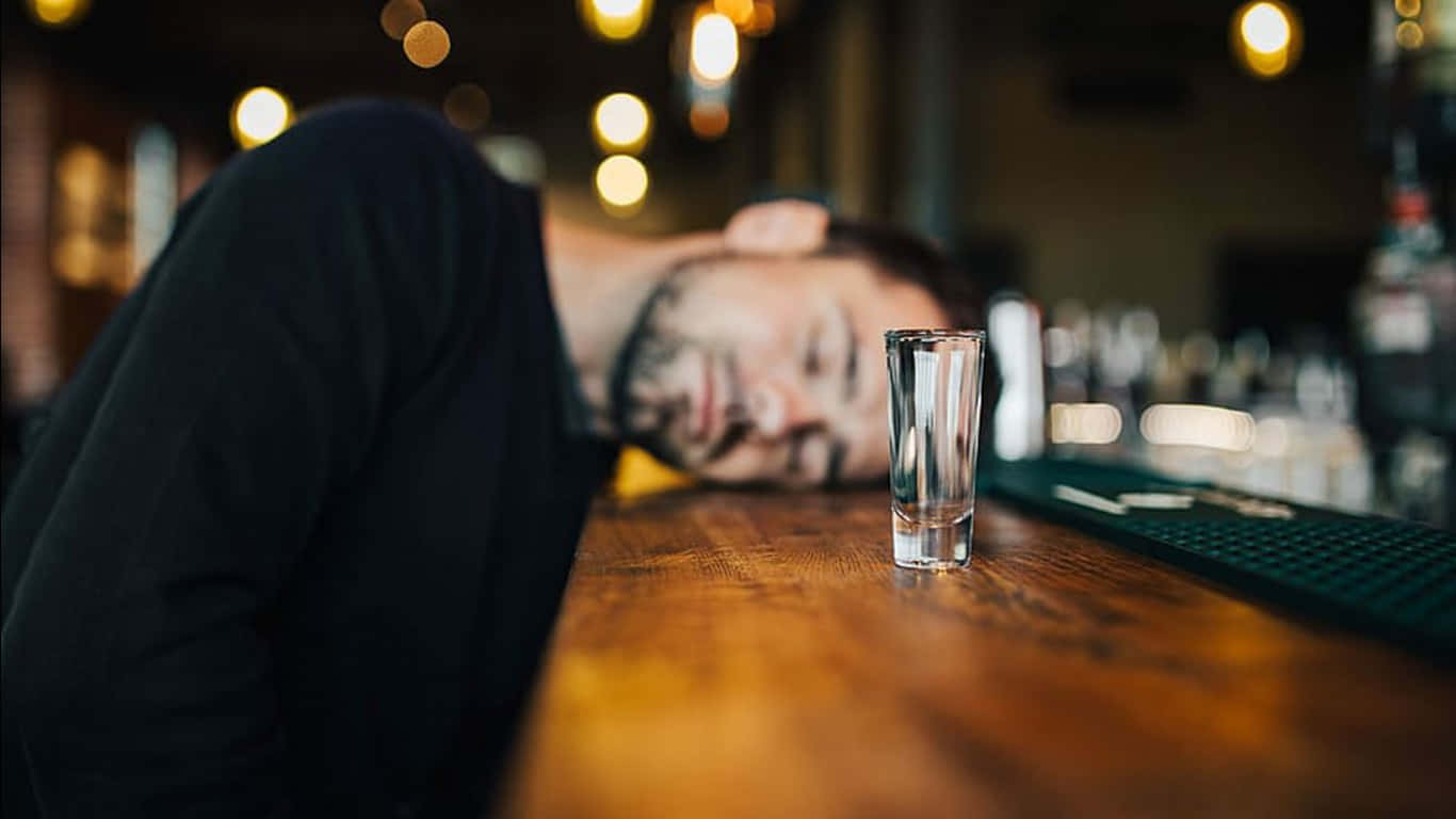  壁紙 — 一名男子在木製吧檯上熟睡，旁邊放著一個空的烈酒杯，溫暖的散景燈光映照下。