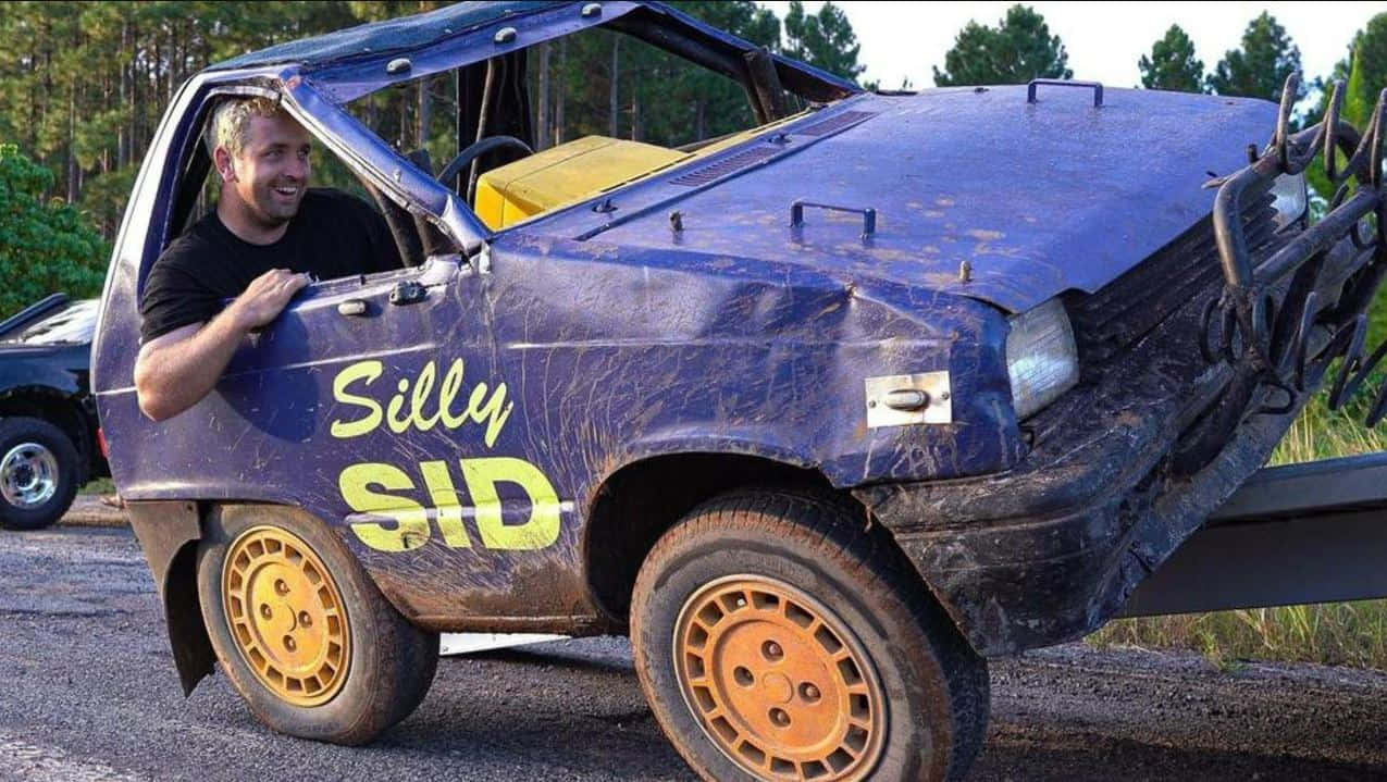  壁紙 — 一名男子在泥濘的藍色越野車Silly Sid的駕駛座上咧嘴笑，車停在森林空地。