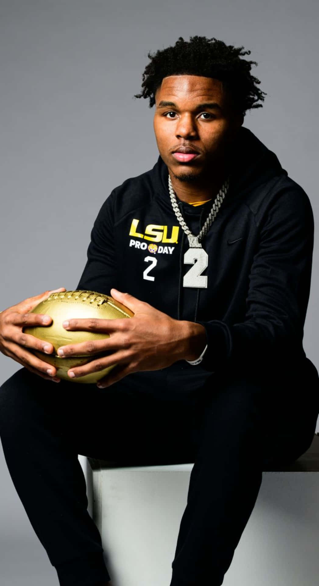  壁紙 — 一名年輕足球員身穿黑色 LSU Pro Day 連帽衫，坐在灰色攝影棚背景前，手持金色足球。