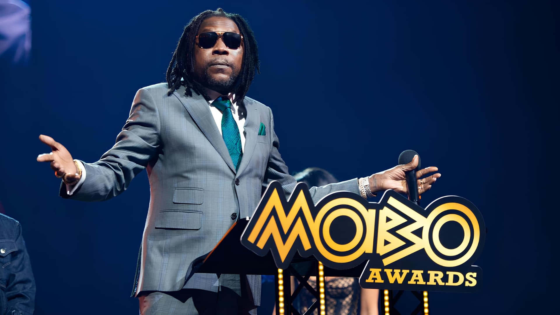  壁紙 — 一名身穿灰色西裝和太陽眼鏡的男子站在MOBO Awards頒獎台上，在藍色舞台燈光下拿著咪高峰。