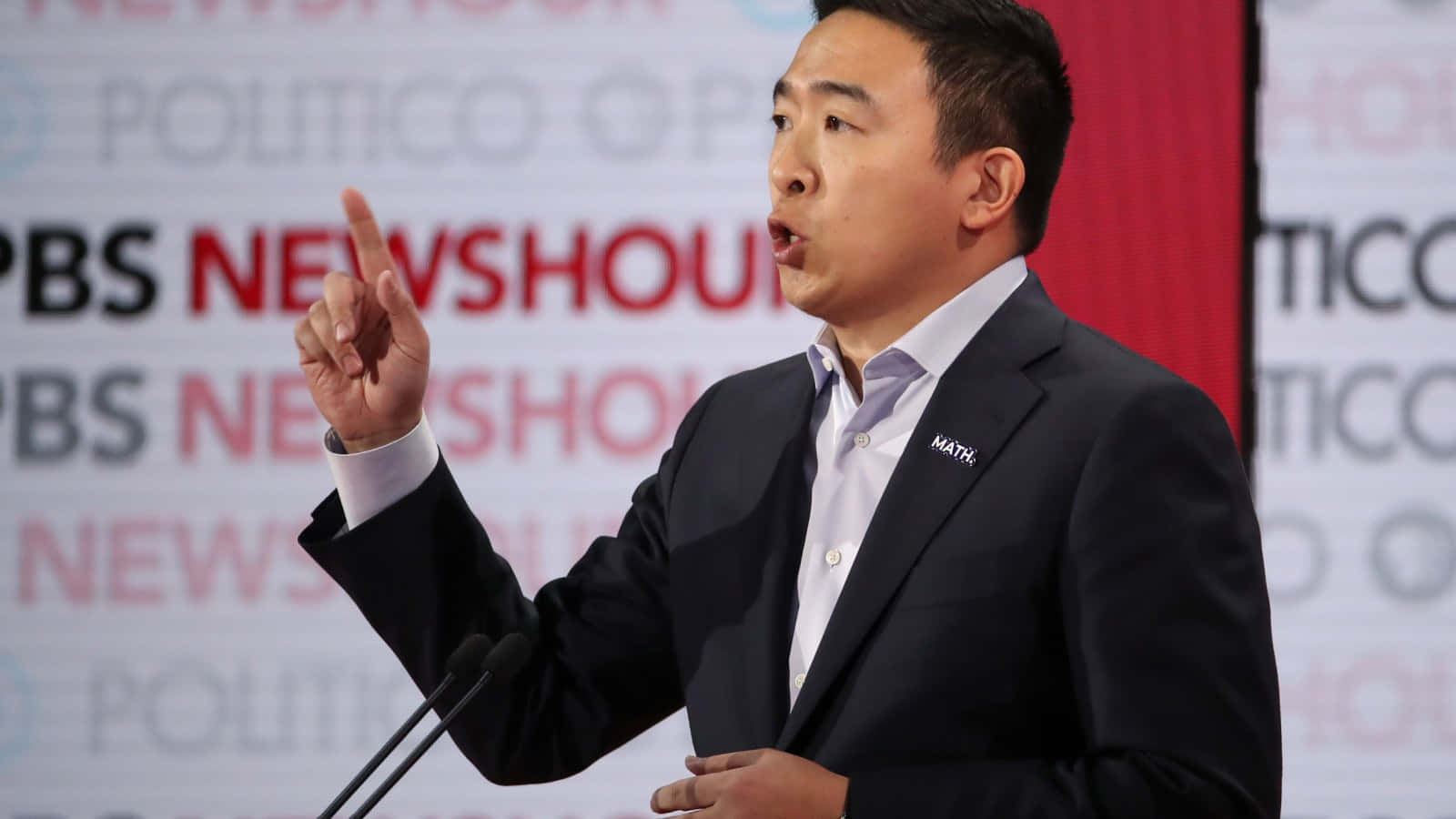 政治 桌布 壁紙 — 一名身穿深色西裝的男子在紅色背景的Politico PBS NewsHour活動中，強調地做手勢並發言。