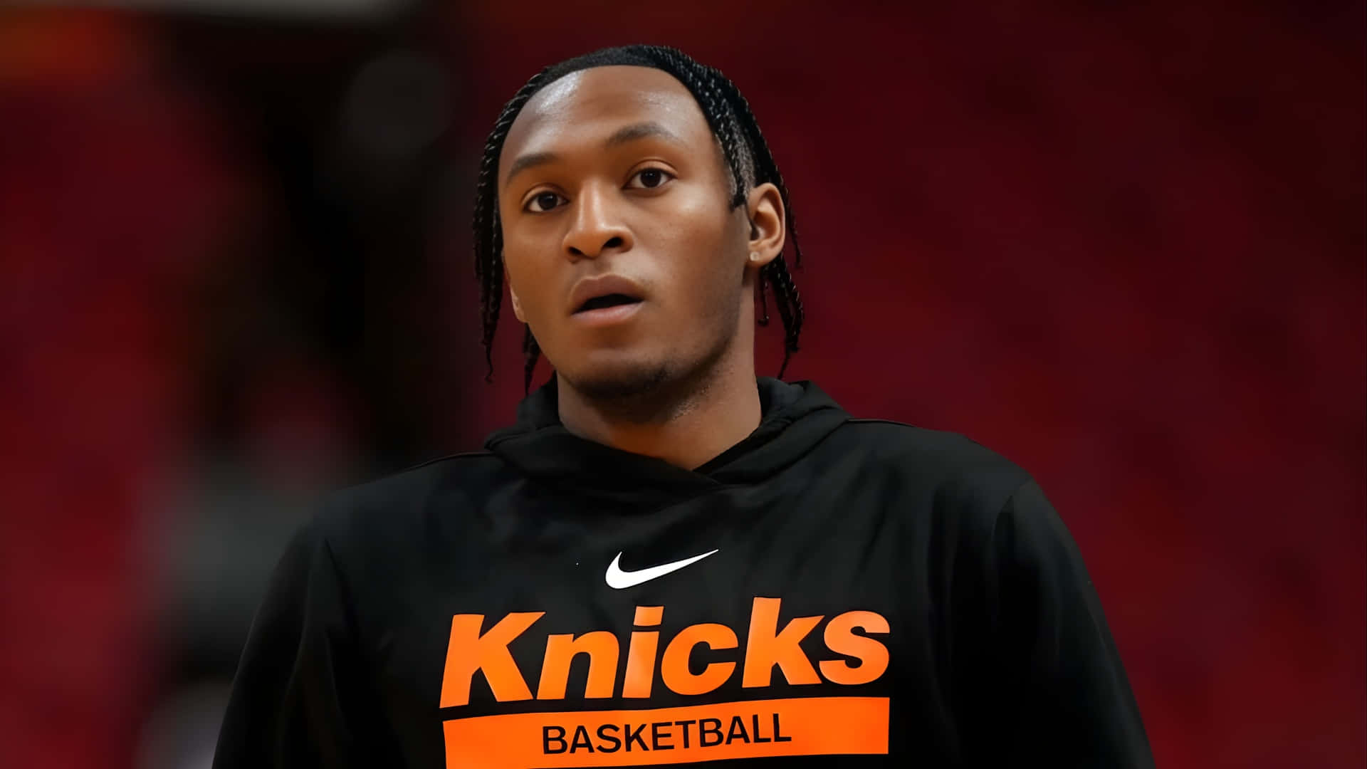 壁紙 — 一名身穿黑色Knicks連帽衫的籃球運動員，在模糊的紅色競技場背景下凝視著。