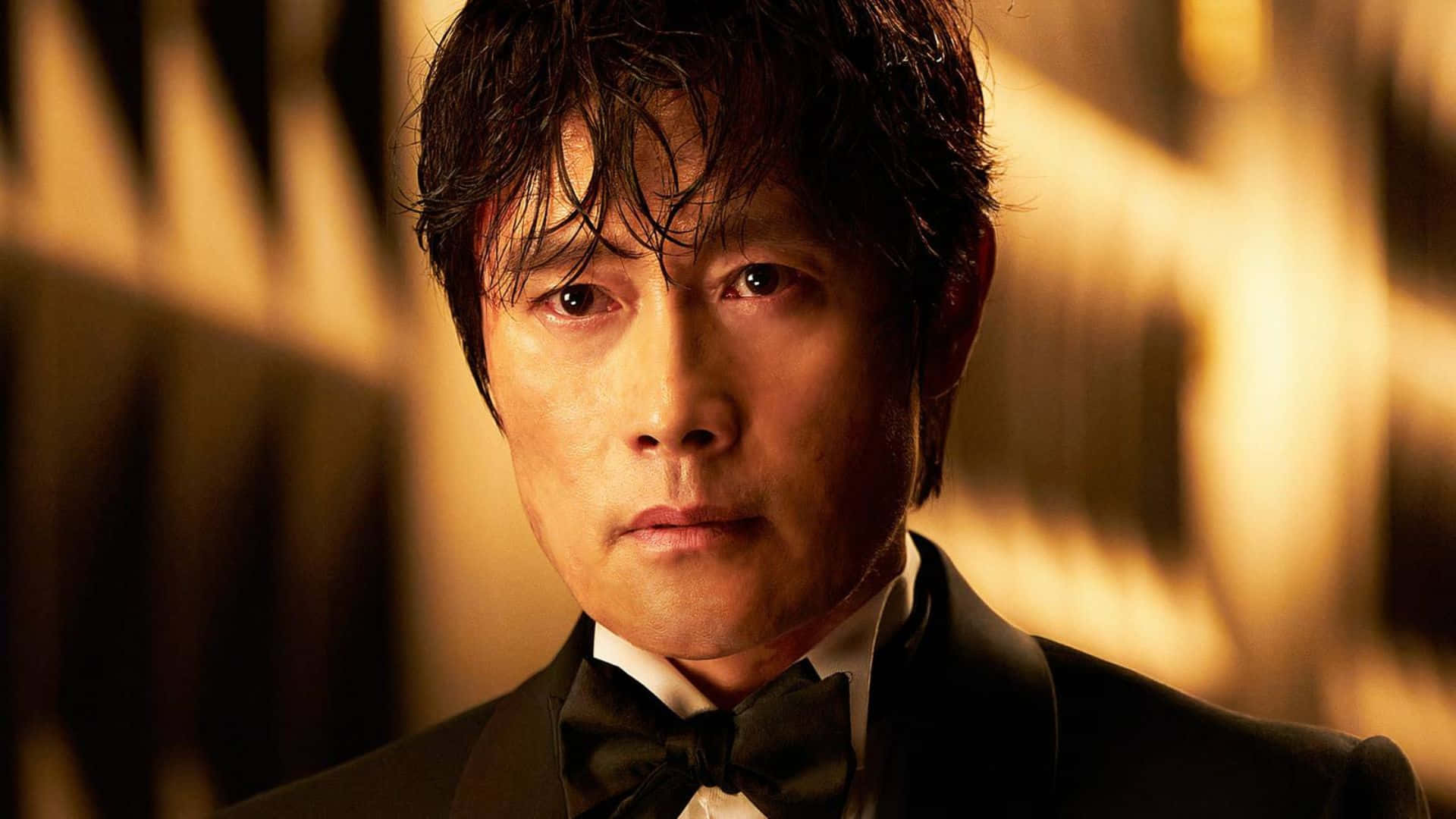  壁紙 — 一名身穿黑色禮服和領結的男子在溫暖的金色燈光下，背景模糊，專注地凝視。