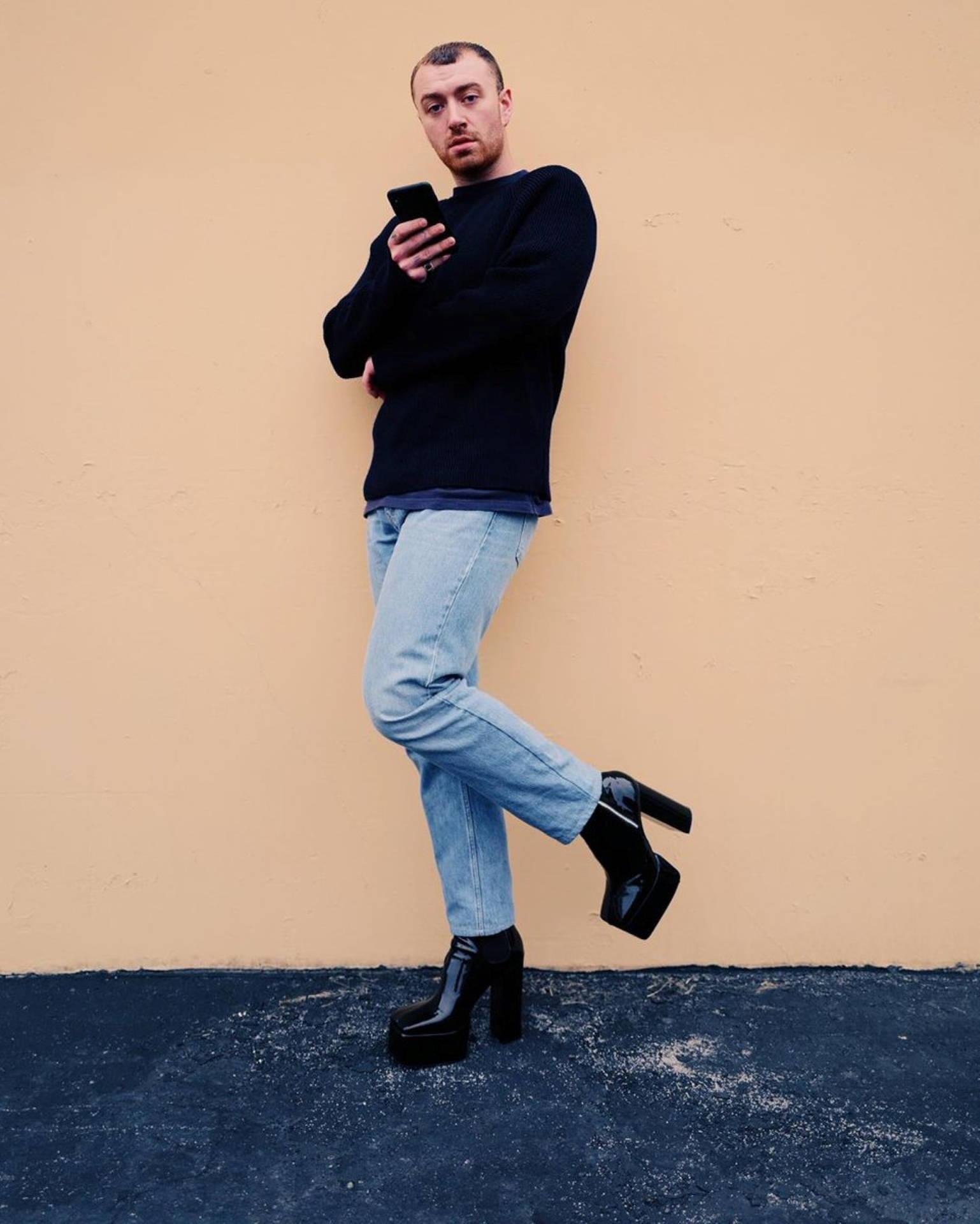 Sam Smith 穿高跟鞋 壁紙 — 一名身穿黑色毛衣和牛仔褲的男子靠著米色牆壁，穿著閃亮的黑色厚底靴，正在看手機。