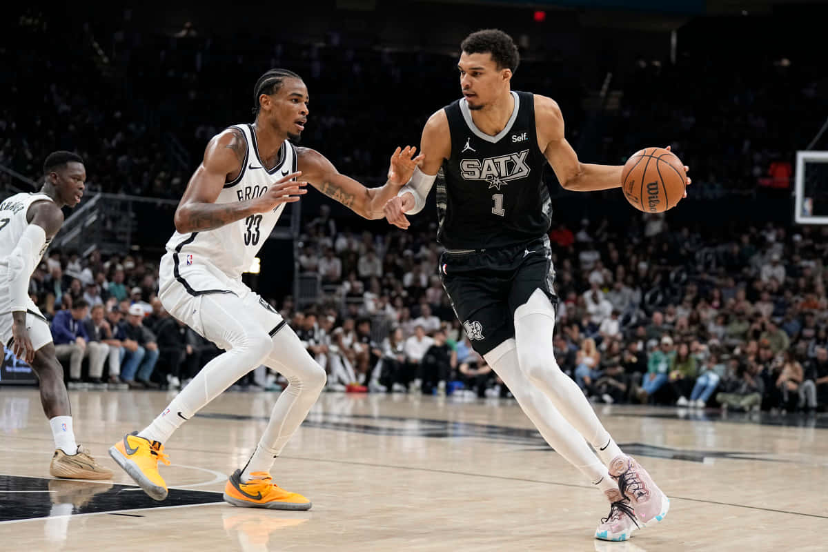  壁紙 — 一名身穿黑色「SATX」球衣的籃球運動員在激烈的比賽中運球，同時被一名身穿白色「BRIO」球衣的對手緊密防守。