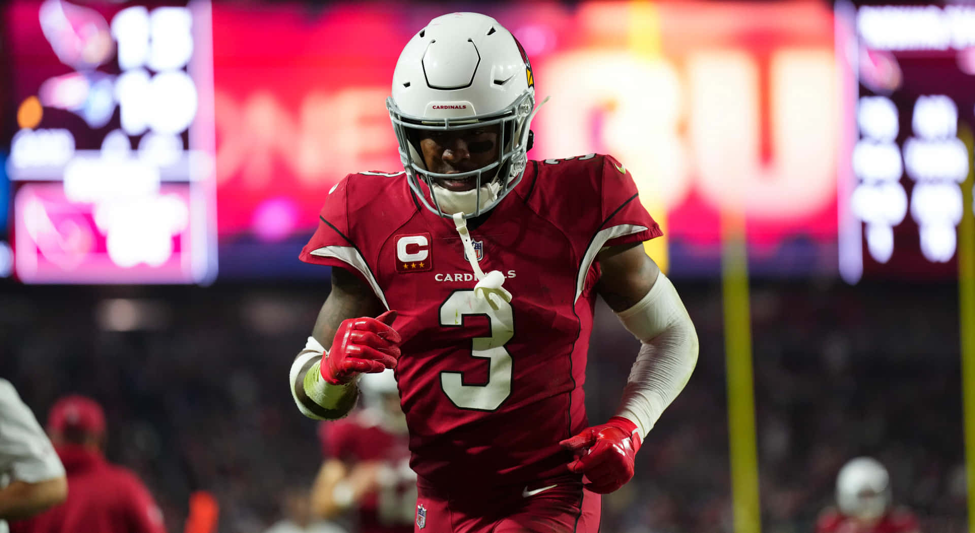  壁紙 — 一名身穿紅色 Cardinals 球衣的美式足球員在明亮的球場燈光下衝過球場，身後有模糊的電子記分板發光。