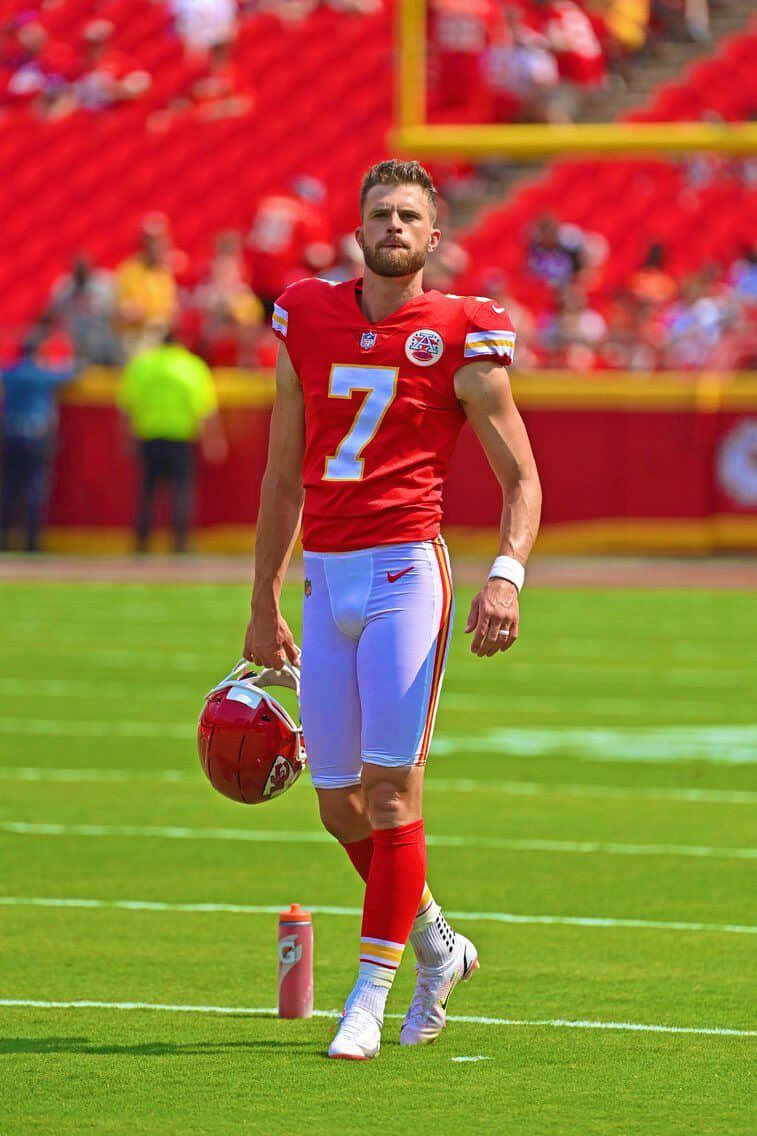 Harrison Butker 桌布 壁紙 — 一名身穿紅色七號球衣的美式足球四分衛在明亮的體育場燈光下拿著頭盔離開球場。