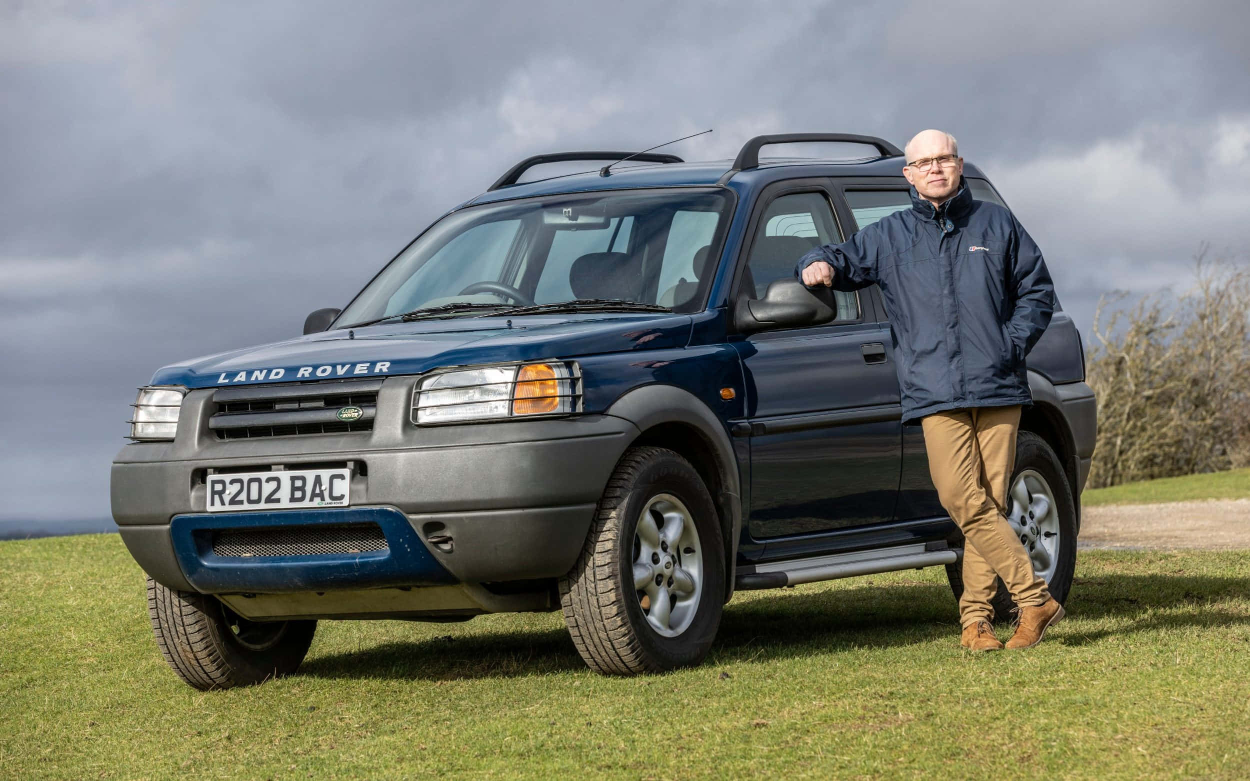 Land Rover Freelander 桌布 壁紙 — 一名身穿藍色外套的男子，在陰天之下，靠在一輛深藍色 Land Rover Freelander 上，背景是長滿草的丘陵。