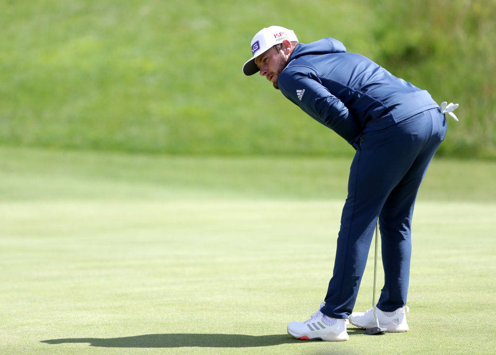 Tyrrell Hatton 桌布 壁紙 — 一名身穿藍色運動服的高爾夫球手在陽光明媚的綠色草地上專注地彎腰準備推桿。