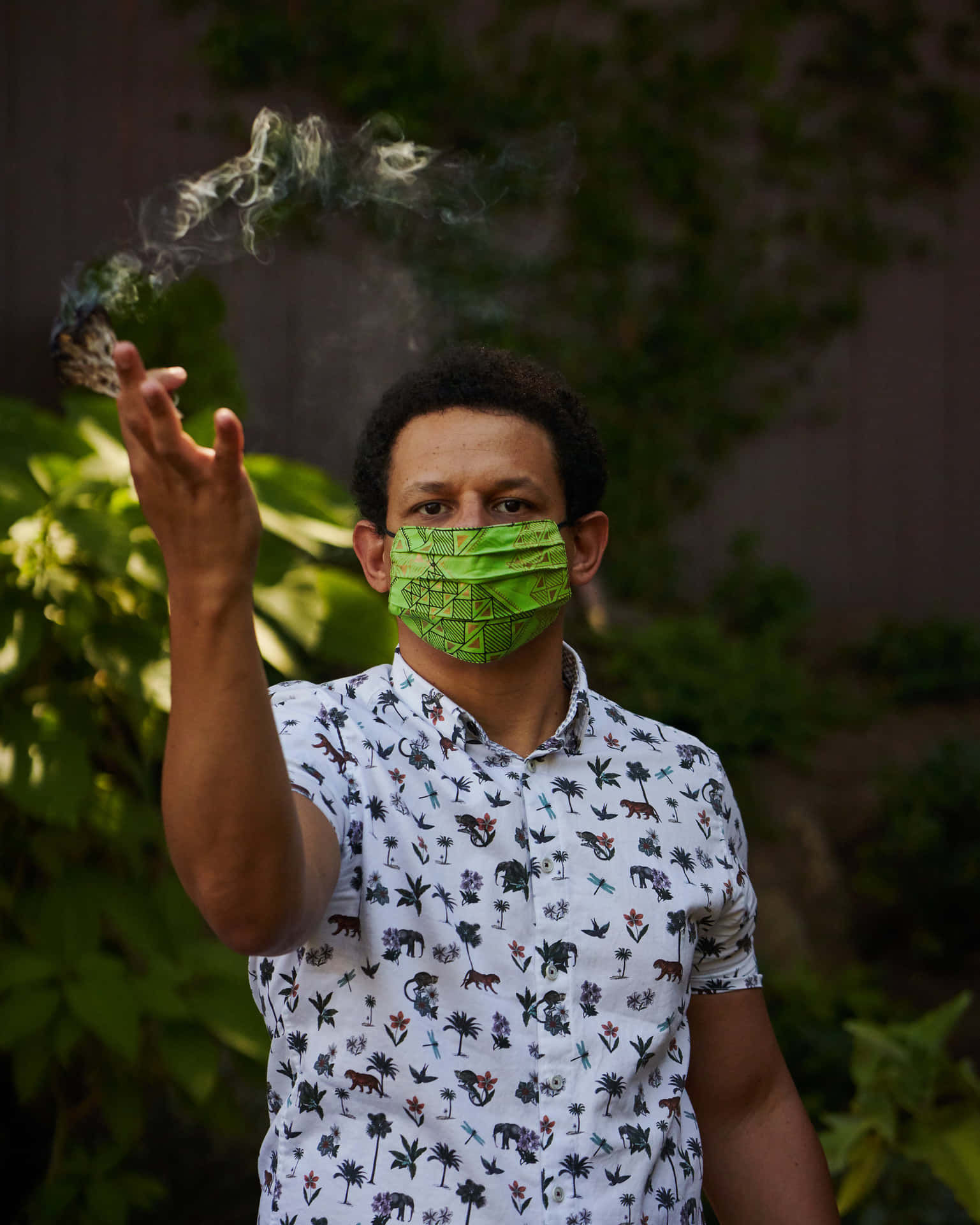  壁紙 — 一名身穿圖案襯衫和綠色口罩的男子在戶外手持冒煙的香煙，身後有茂密的綠植。