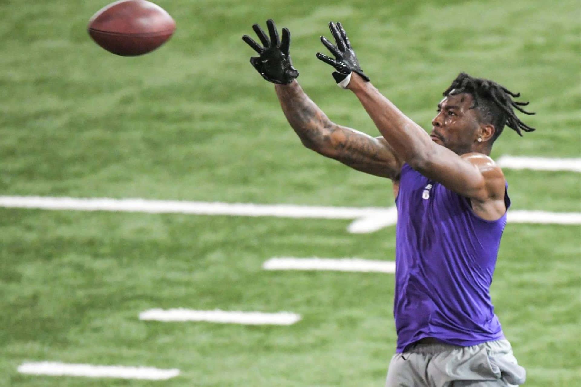  壁紙 — 一名身穿紫色球衣的足球運動員戴著手套跳起，在綠色球場上伸手接一個棕色足球。