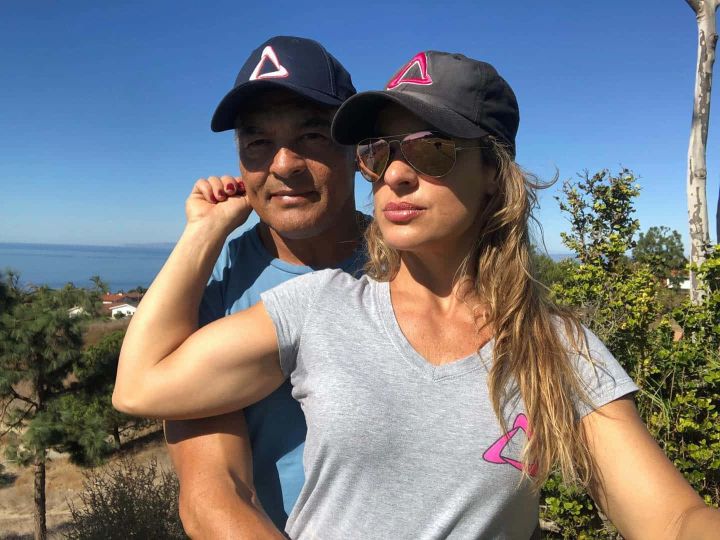  壁紙 — 一男一女戴著相配的帽子，在陽光明媚的山坡上自拍，俯瞰海洋和茂密的綠植。