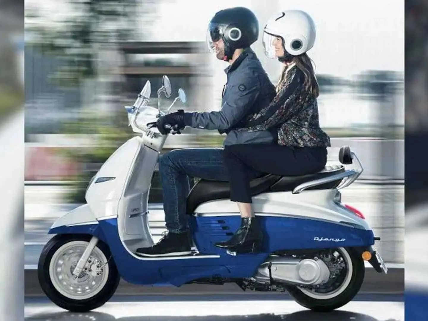 Peugeot Motorcycle 桌布 壁紙 — 一男一女戴著頭盔，騎著藍白相間的電單車行駛在模糊的城市街道上。