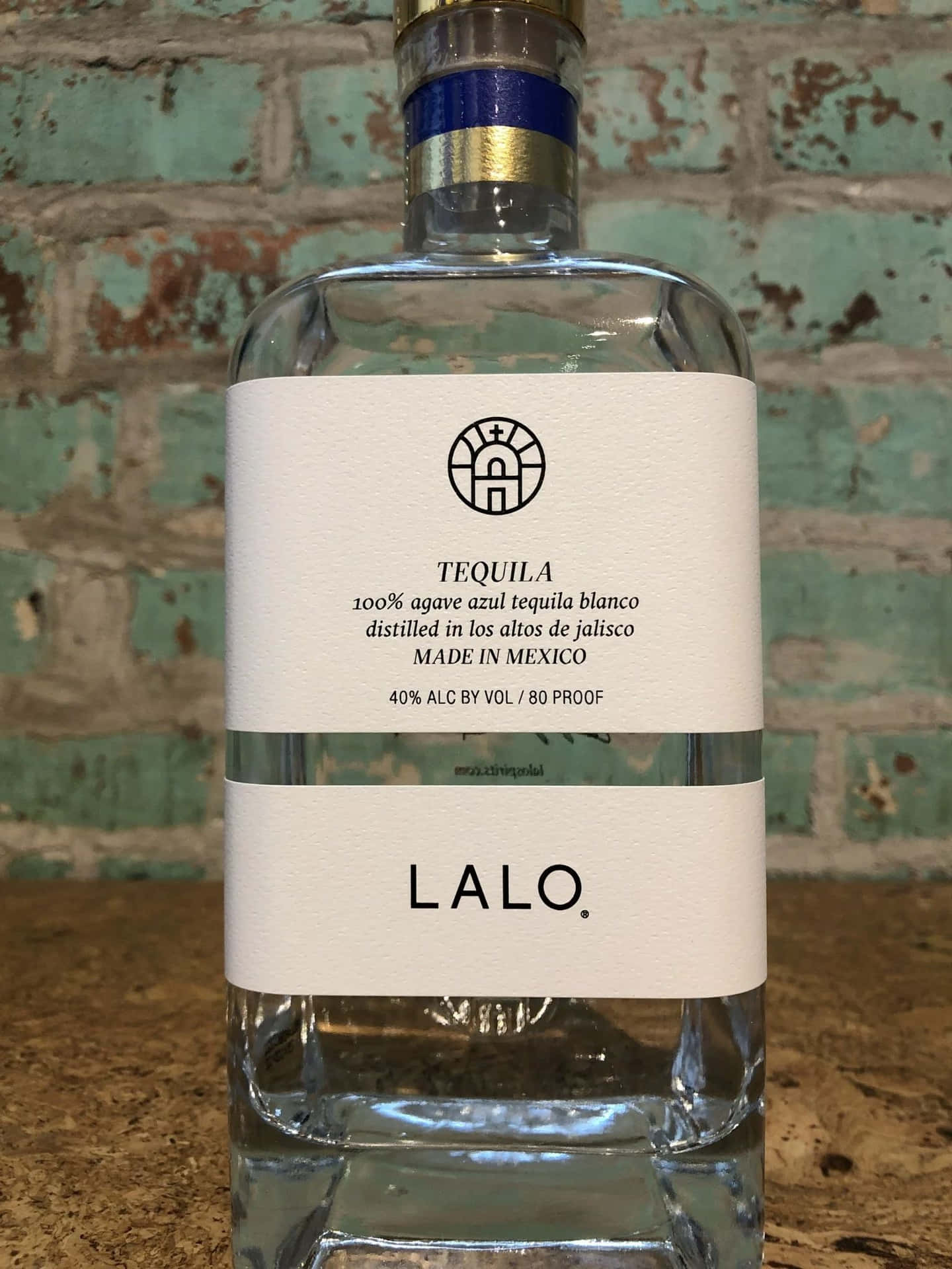  壁紙 — 一瓶清澈的Lalo龍舌蘭酒，靠著一面風化的青綠色磚牆，散發質樸魅力。