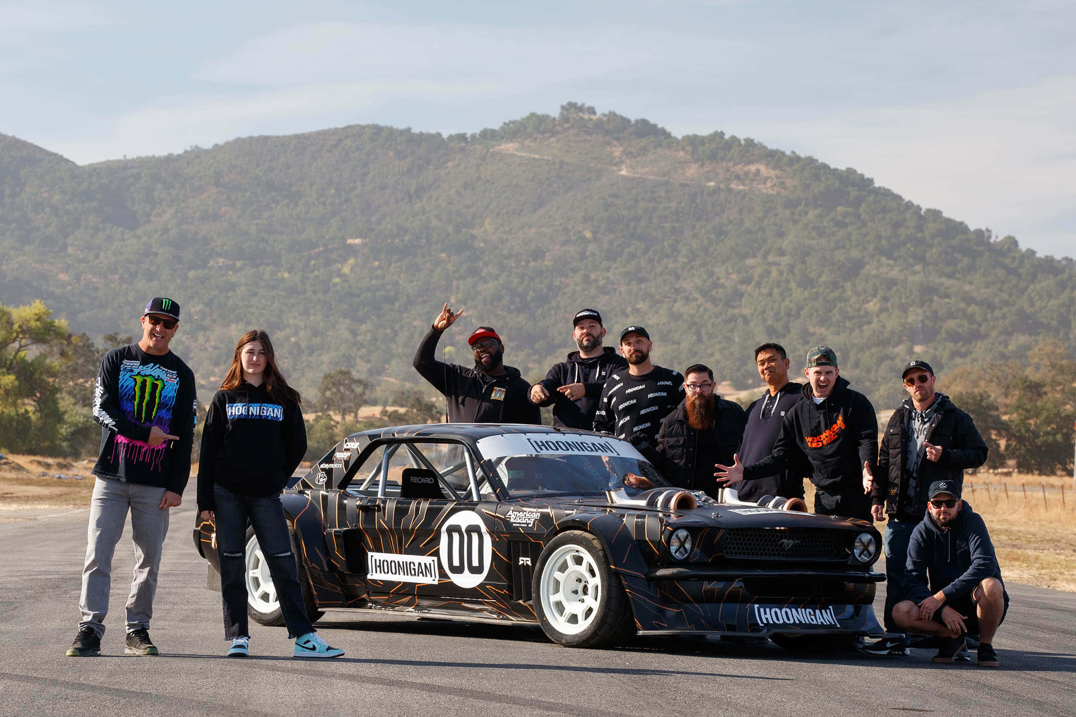 Ken Block 桌布 壁紙 — 一群人自豪地站在一輛帶有橙色點綴的黑色Hoonigan賽車旁，背景是鋪砌的道路和起伏的山丘。
